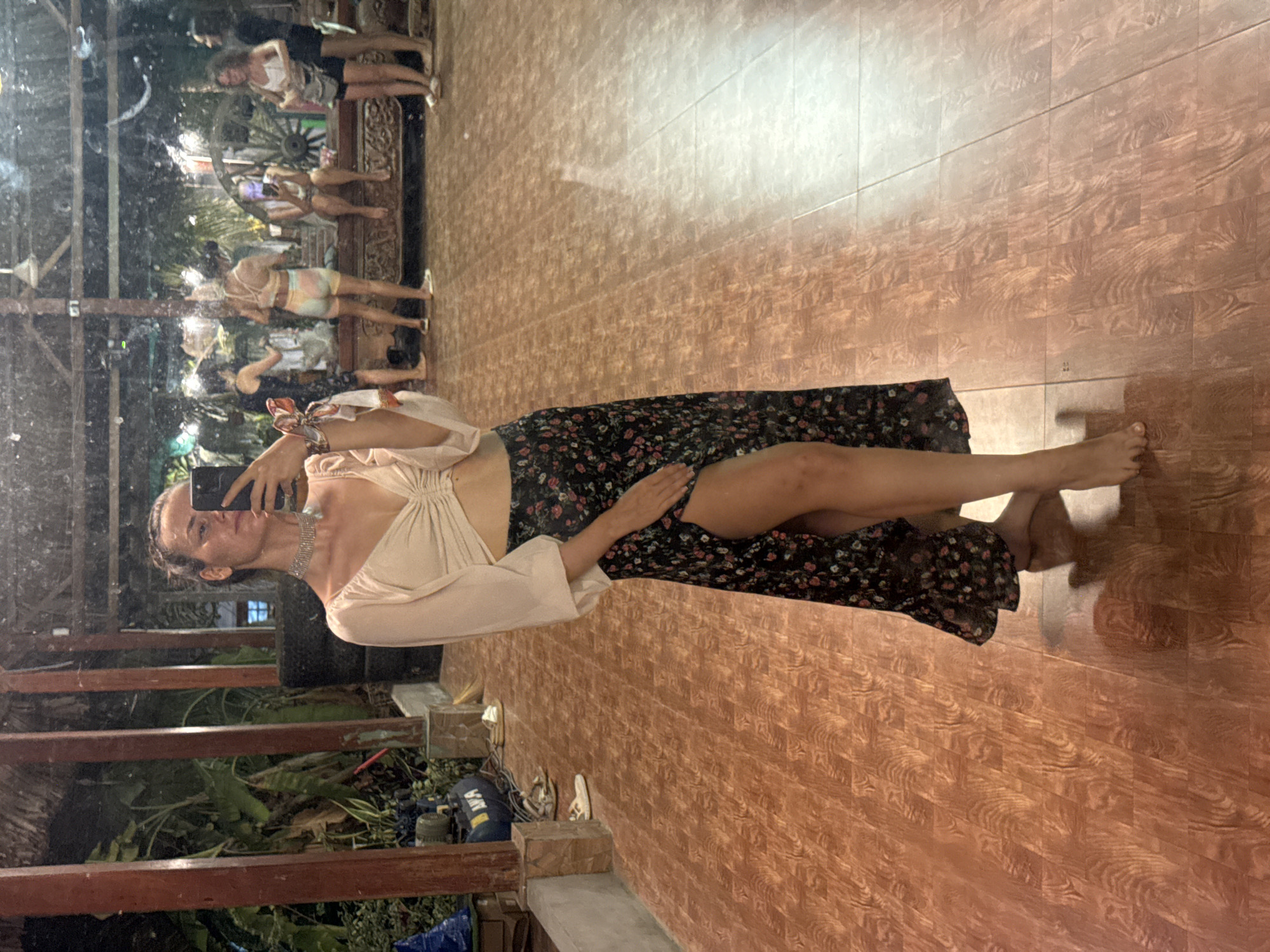 A woman stands facing a mirror, holding a smartphone and taking a selfie. She wears a light-colored top and a dark, floral-patterned skirt with a slit. The reflection in the mirror shows a blurred scene of people and objects. The environment appears to be an indoor-outdoor space with tiled flooring. Outside the mirror, there's greenery and some equipment. The location is Phu Thuy Ward, Vietnam.