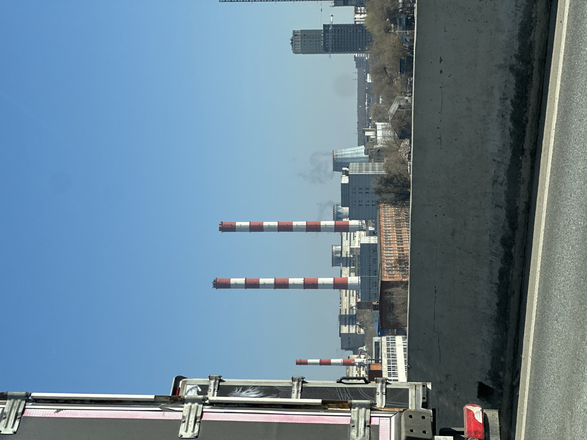 Here is a description of the image:
The image shows a view from what appears to be a vehicle on a bridge or overpass, overlooking a cityscape. Prominent features include three tall industrial smokestacks with alternating red and white stripes, which are characteristic of the Moscow heating plants. The smokestacks are positioned centrally in the frame, rising above a cluster of buildings, indicating an industrial or urban setting. In the background, there are taller buildings and high-rises, suggesting a city center. The sky is clear blue, providing a bright backdrop. The foreground includes the edge of the bridge and some structures or equipment likely attached to the vehicle from which the photo was taken. Based on the smokestacks' distinctive coloring and design, and the overall urban landscape, the location is likely in Moscow, Russia.