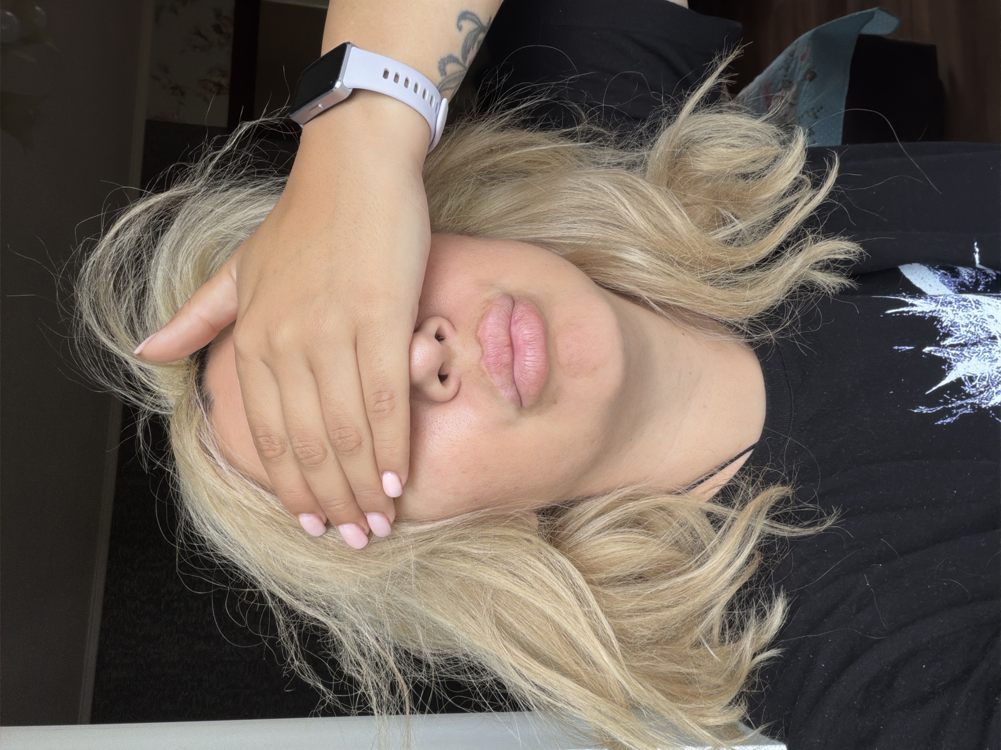 A woman with blonde hair is covering her eyes with her right hand. Her mouth is pursed, and she is wearing a black t-shirt and a light-colored smartwatch. The background appears to be an indoor setting with a dark wall and a window. There is no discernible location in the city of Partenit, Ukraine.