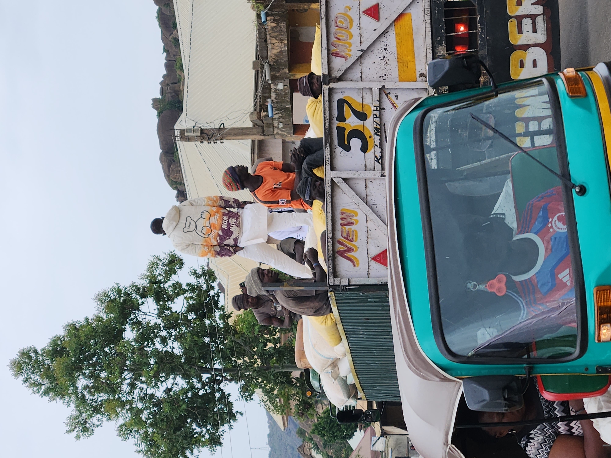 In Rukuba Road, Nigeria, a flatbed truck is loaded with numerous large, pale yellow sacks, likely agricultural produce. Several individuals are positioned atop these sacks and on the truck's railings. One man in the foreground wears an orange t-shirt and a knitted cap, while another to his right is dressed in a cream-colored hoodie adorned with a cartoon bear and orange and purple graphics, paired with white trousers. A third person, wearing a dark beanie, sits on the truck's edge to the left of the man in the orange shirt. The truck's side displays faded white paint with painted text including "NEW," "57," and "MOD." Two other men are visible behind the front figures. In the foreground, a teal-colored auto-rickshaw is partially visible, with a person in the driver's seat wearing a blue and red striped shirt. The background reveals a tree with dense green foliage and a rocky, elevated terrain with some buildings. The sky is overcast.