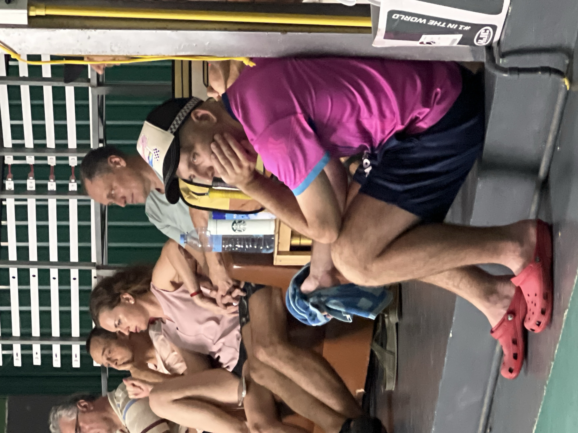 A group of individuals are seated on tiered seating, possibly at a sporting event or performance, within a building. The scene is partially illuminated, with the background featuring vertical white and green slatted structures.
In the foreground, a man wearing a bright pink t-shirt and dark blue shorts is seated and leaning against a gray concrete wall. He has a black and white checkered cap on his head and is resting his chin on his hand. In front of him, to his left, are a pair of red Croc-style shoes.
To his left, a woman in a light pink sleeveless top is seated, with her legs crossed and bare. She is looking to her left, with her left hand resting on her knee. Next to her, a man in a light colored t-shirt is also seated, looking to his left with his hand near his face. Another man is partially visible on the far left, wearing glasses and a striped t-shirt.
Between the seated individuals are a wooden crate and two plastic water bottles. A blue and white towel is draped over the armrest of the seating.
The location is identified as Ban Bang Rak, Thailand. The overall environment suggests an indoor venue with spectator seating.