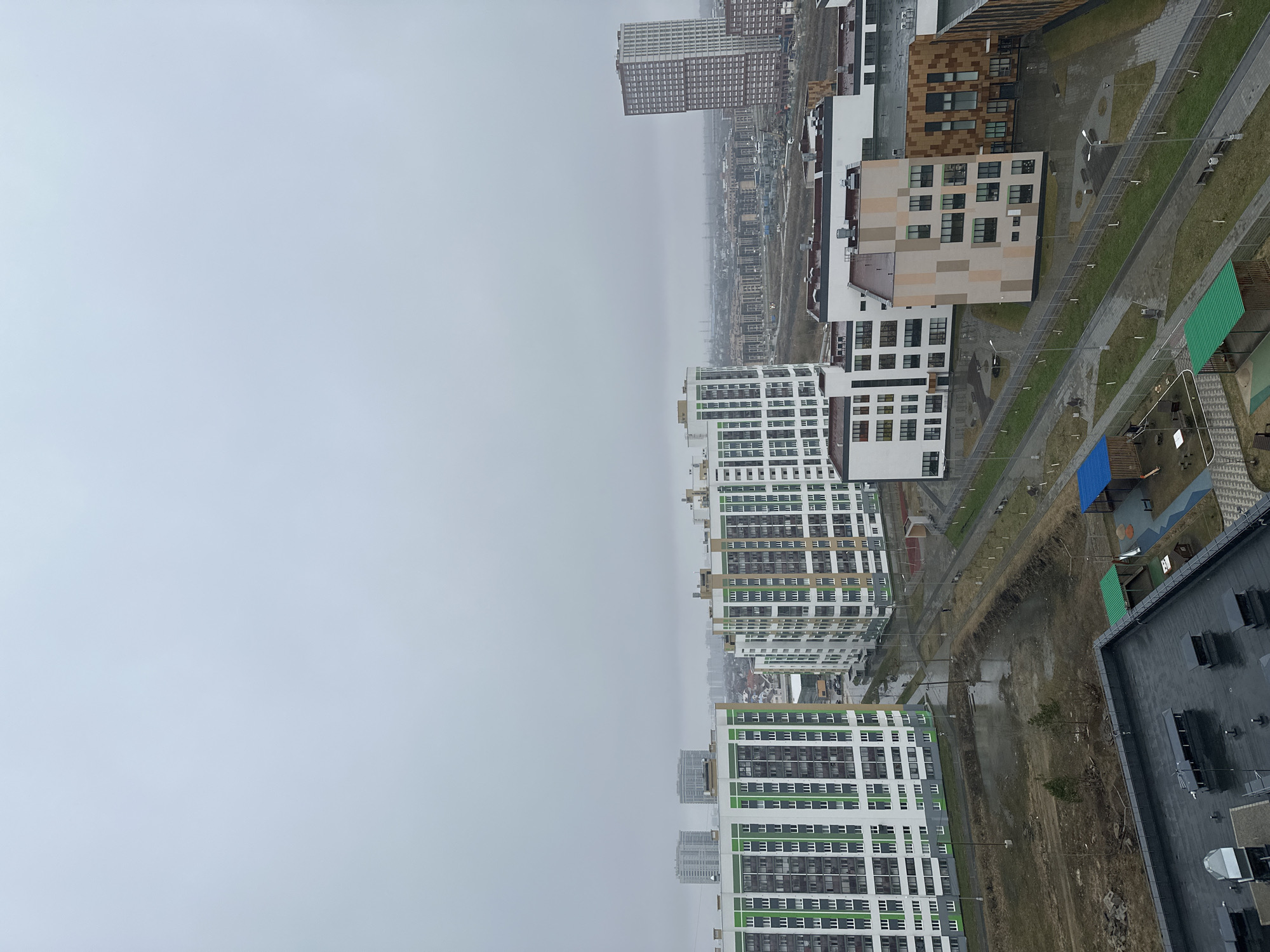This high-angle, panoramic view captures a modern urban landscape in Yekaterinburg, Russia, under a heavily overcast sky. The image appears to be taken from a tall building, looking out over a sprawling residential development.
The scene is dominated by numerous high-rise apartment buildings. In the midground and foreground, several contemporary buildings feature distinct white facades with prominent green accents, arranged in horizontal and vertical patterns, and countless windows. Other buildings display a mosaic of beige and brown panels, suggesting varied architectural styles within the development. Further in the distance, additional high-rise structures, appearing slightly older and more uniform in their grey tones, stretch towards a hazy horizon, indicative of a large urban sprawl.
At ground level, a network of paved pathways and roads meanders through grassy areas. Many sections of grass appear sparse, light green, or even muddy, suggesting recent construction or ongoing landscaping. Modern streetlights with multiple arms line these paths. A children's playground area, distinguished by sections covered in bright blue and green material, is visible. In the bottom right corner, a large building with a flat, dark roof and visible utility units stands, possibly a commercial or industrial structure.
The weather is characterized by a uniform, dull grey overcast sky that occupies a significant portion of the upper and left side of the frame, giving the entire scene a muted and cool appearance. Surfaces like pavements and roofs appear damp, hinting at recent rain or persistent damp conditions. No people or specific activities are visible, emphasizing the expansive and somewhat stark nature of the newly developed area. There is no discernible text visible on any of the buildings or objects.