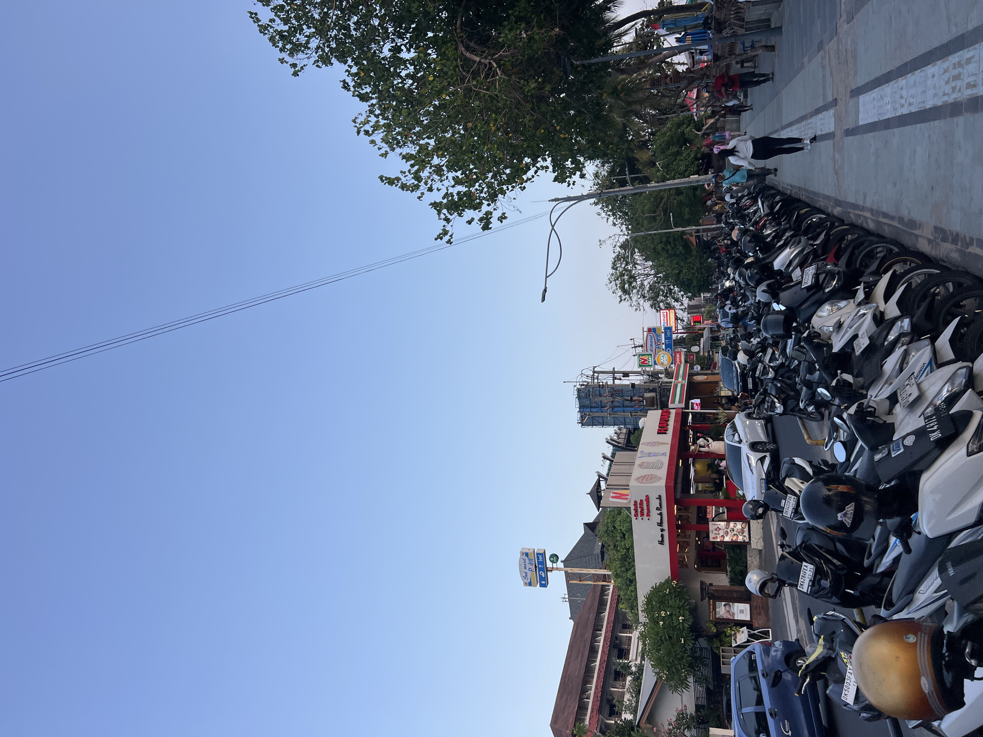 The image depicts a street scene in Kuta, Indonesia, characterized by a clear blue sky and a line of parked motorcycles. The motorcycles are primarily lined up along a paved sidewalk, creating a dense parking area. Several of these motorcycles have helmets placed on them. To the left of the motorcycles, a road is visible with a blue car and a white SUV driving on it. Further down the street, various buildings are present, including a store with a prominent red and white awning displaying the text "House of Banana Pancake" and an "Indomaret POINT" sign. Trees with green foliage are also scattered throughout the scene, some overhanging the sidewalk. A few individuals are walking on the sidewalk, and others are seen near the storefronts. Power lines are visible crisscrossing the upper portion of the sky. The overall atmosphere suggests a busy commercial area, possibly a tourist destination, with a significant presence of two-wheeled transportation.