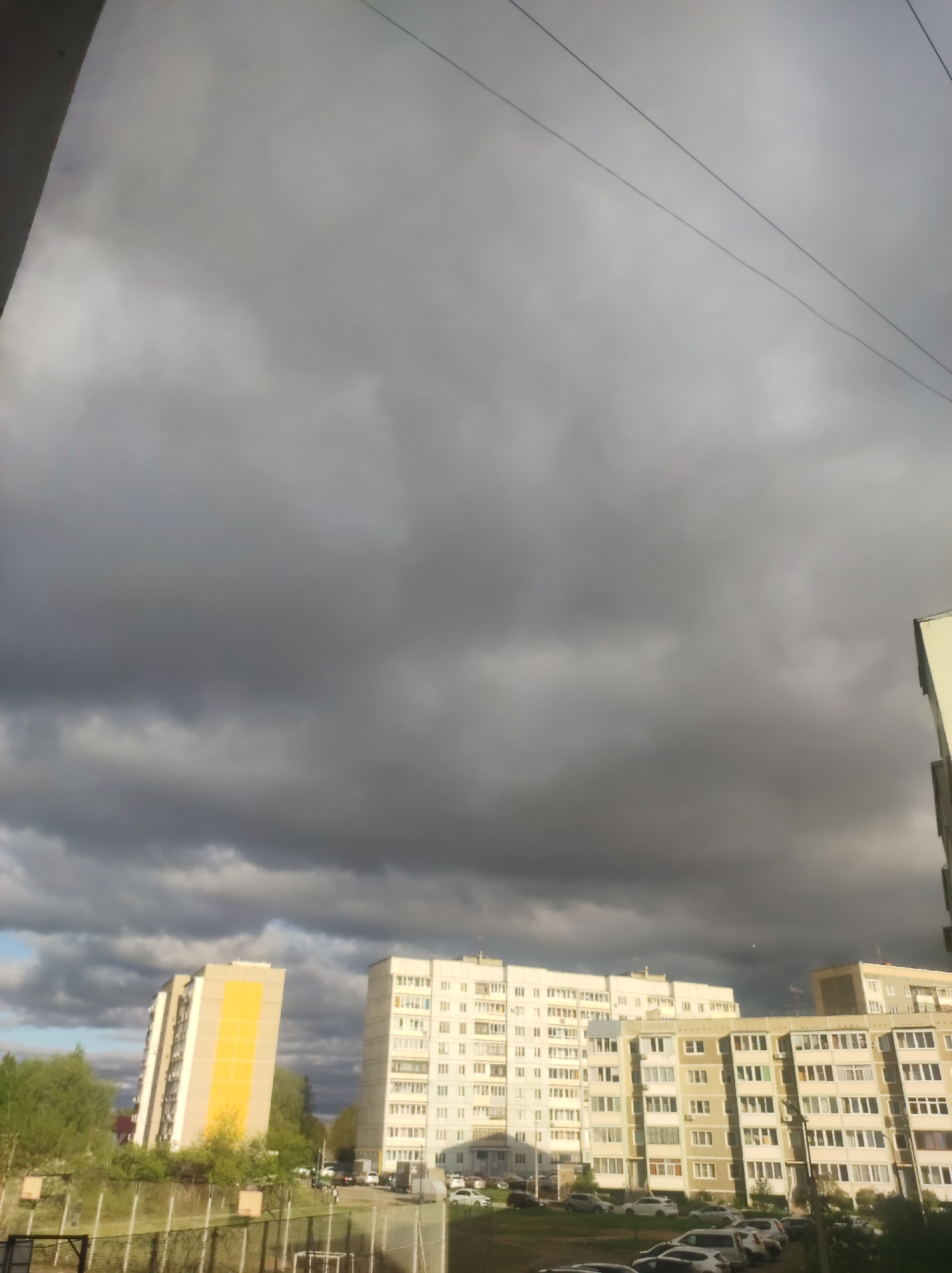 The photograph depicts an urban scene in Chaikovsky, Russia. Several multi-story apartment buildings dominate the midground. The sky above is overcast with heavy, dark storm clouds. Power lines run across the upper part of the frame. Fencing surrounds vegetation in the foreground. Parked cars are visible near the apartment buildings. No people are clearly visible.