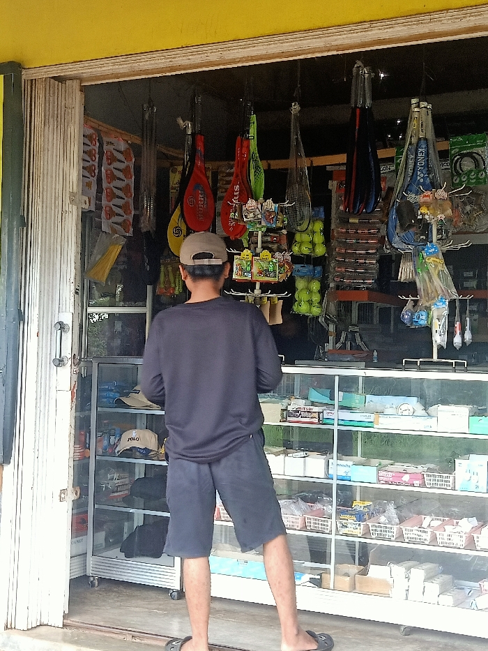 The image captures a man standing, viewed from behind, at the entrance of a small shop in Pubian, Indonesia. The shop appears to be a local general store, possibly specializing in sports equipment, with a yellow-painted facade above the entrance.

The man is wearing a dark long-sleeved shirt, dark shorts, and a beige baseball cap. He is also wearing flip-flops. His posture suggests he is looking into the shop, possibly browsing the merchandise or engaging with someone inside.

Inside the brightly lit shop, a variety of items are displayed. Hanging from the ceiling are numerous badminton rackets in various colors, including red, yellow, green, and blue. Some rackets clearly show brand names like "SPORT" and "YONEX". Beside these, several mesh bags filled with yellow tennis balls are visible. Other hanging items include what appear to be fishing nets and packaged scissors.

The shop's interior features glass display cases with multiple shelves, holding a diverse range of products. A white baseball cap with the word "POLO" is visible on a lower shelf in one of the display cases to the left. Small items are arranged on a rotating display stand, and various boxes and baskets of goods fill the shelves. The shop entrance is flanked by a white corrugated rolling shutter, partially open on the left. The scene depicts a typical daytime street-side shopping experience in a local setting.
