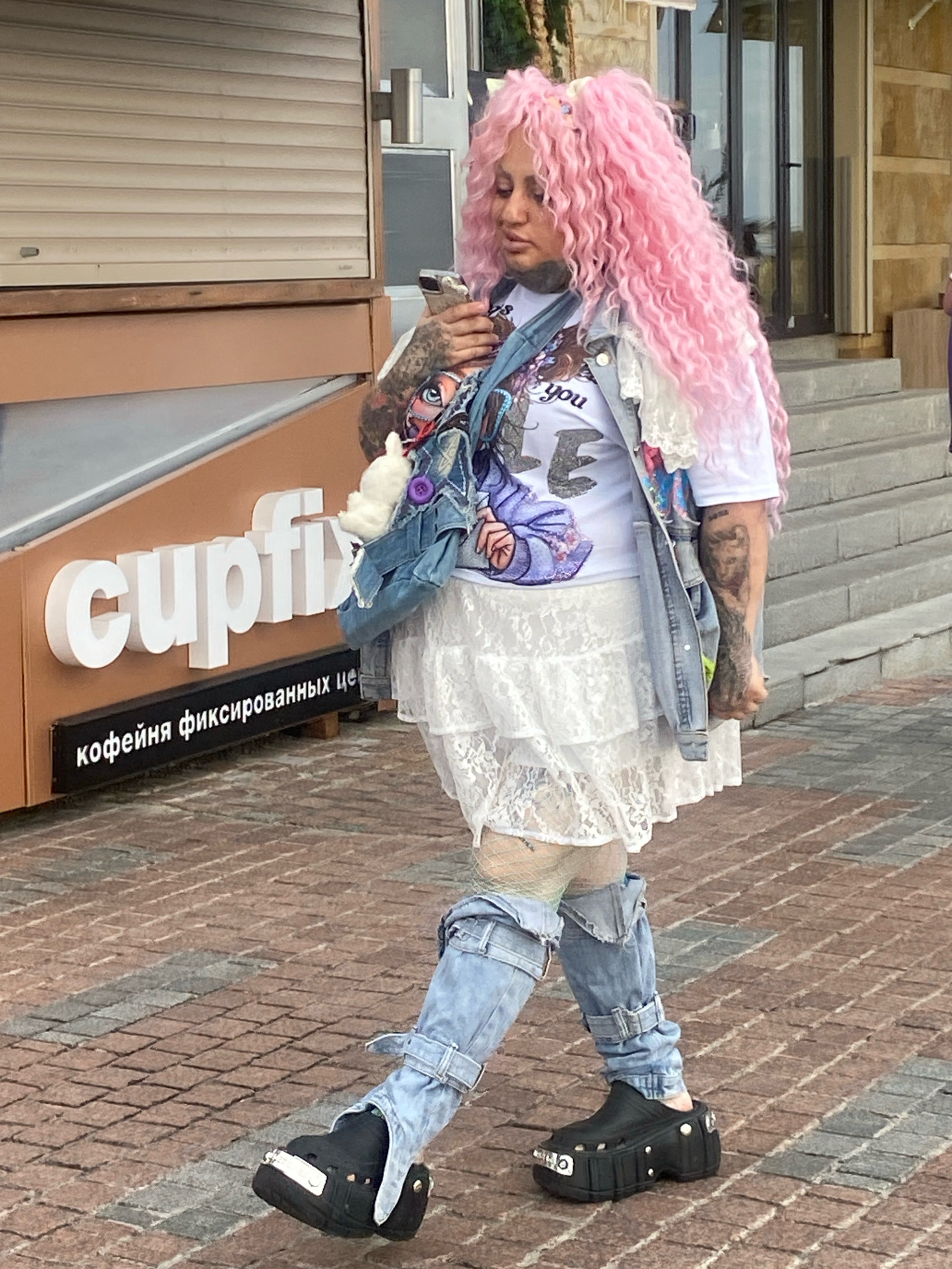 A woman with bright pink curly hair walks past a storefront in Sochi, Russia. She is wearing a white t-shirt with a graphic print, a white lace skirt, and distressed denim jeans. She also has on black platform clogs and a denim bag with a white stuffed animal attached. The storefront has a prominent sign with the word "cupfix" in large white letters, and below it, in Russian, "кофейня фиксированных цен" which translates to "coffee shop with fixed prices." The ground is paved with brick.