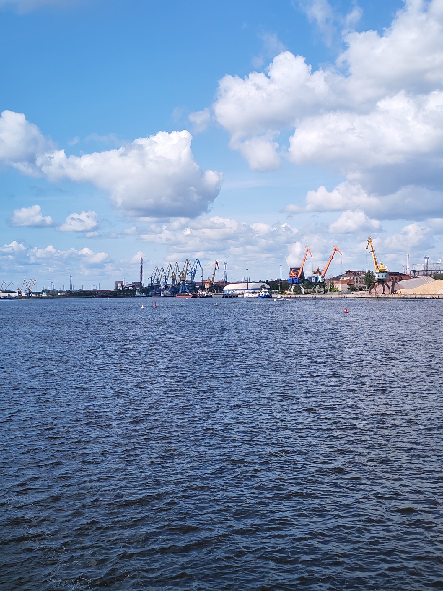 The image depicts the Cherepovets port on the Rybinskoye Reservoir, with a view of the northern shoreline. The foreground is dominated by dark blue, choppy water. In the background, the port facilities are visible under a bright blue sky with scattered white clouds. A line of various industrial cranes, including tower cranes and gantry cranes, stretches along the waterfront. Some of these cranes are orange, blue, and yellow. Several buildings, including industrial structures and what appear to be warehouses, are situated behind the cranes. Piles of sand or aggregate are visible near some of the facilities. A few small boats or barges are present in the water near the shore. The overall scene is one of an active industrial port.