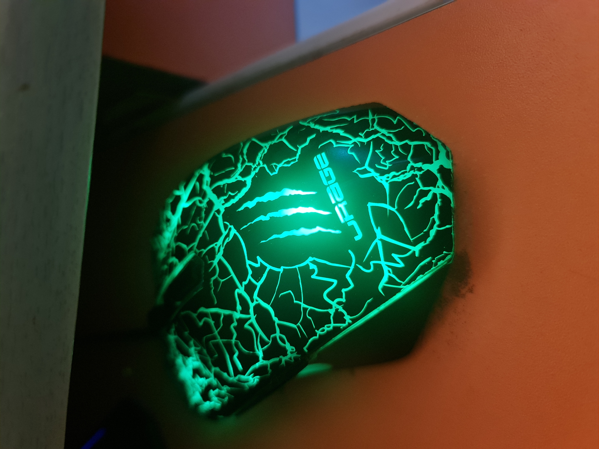 The image captures a close-up of a gaming mouse, illuminated with a vibrant green light, in a dimly lit indoor setting. The mouse, predominantly black, features a striking "cracked" or "lightning bolt" pattern across its surface, which glows intensely green. On its top, the brand name "URAGE" is visible in a stylized font, also glowing green, positioned above a bright green claw mark design.
The mouse is situated against an orange-textured surface, likely a wall or furniture, which subtly reflects some of the green light. To the left, a light-colored vertical element, possibly a door frame, partition, or furniture edge, partially obscures the background. Above the mouse, a narrow, light-colored horizontal strip suggests a shelf or trim. The overall scene indicates a low-light environment, such as a room at night or a dedicated gaming setup, emphasizing the mouse's illuminated features. No discernible human activity or specific event is taking place beyond the mouse being powered on. The location context provided is Kazanlak, Bulgaria.