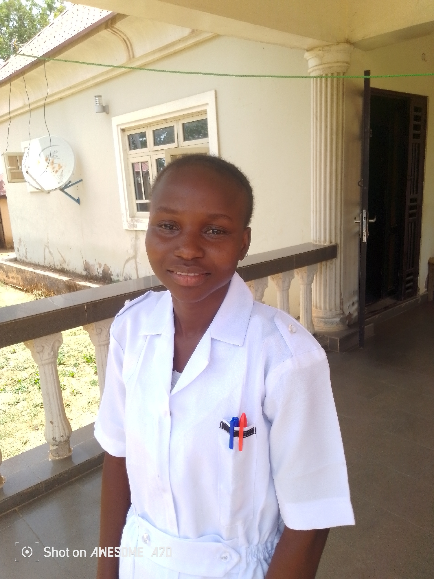A medium shot captures a young Black woman in a white uniform, presumably a nurse or healthcare worker, smiling gently at the camera. Two pens, one red and one blue, are clipped to the pocket of her uniform.
She stands on a veranda with a low, decorative balustrade. Behind her, the building has white walls, a satellite dish is attached. The doorway is open to a dark interior.
The location is listed as Unknown, Unknown. The environment appears to be a residential or medical setting.