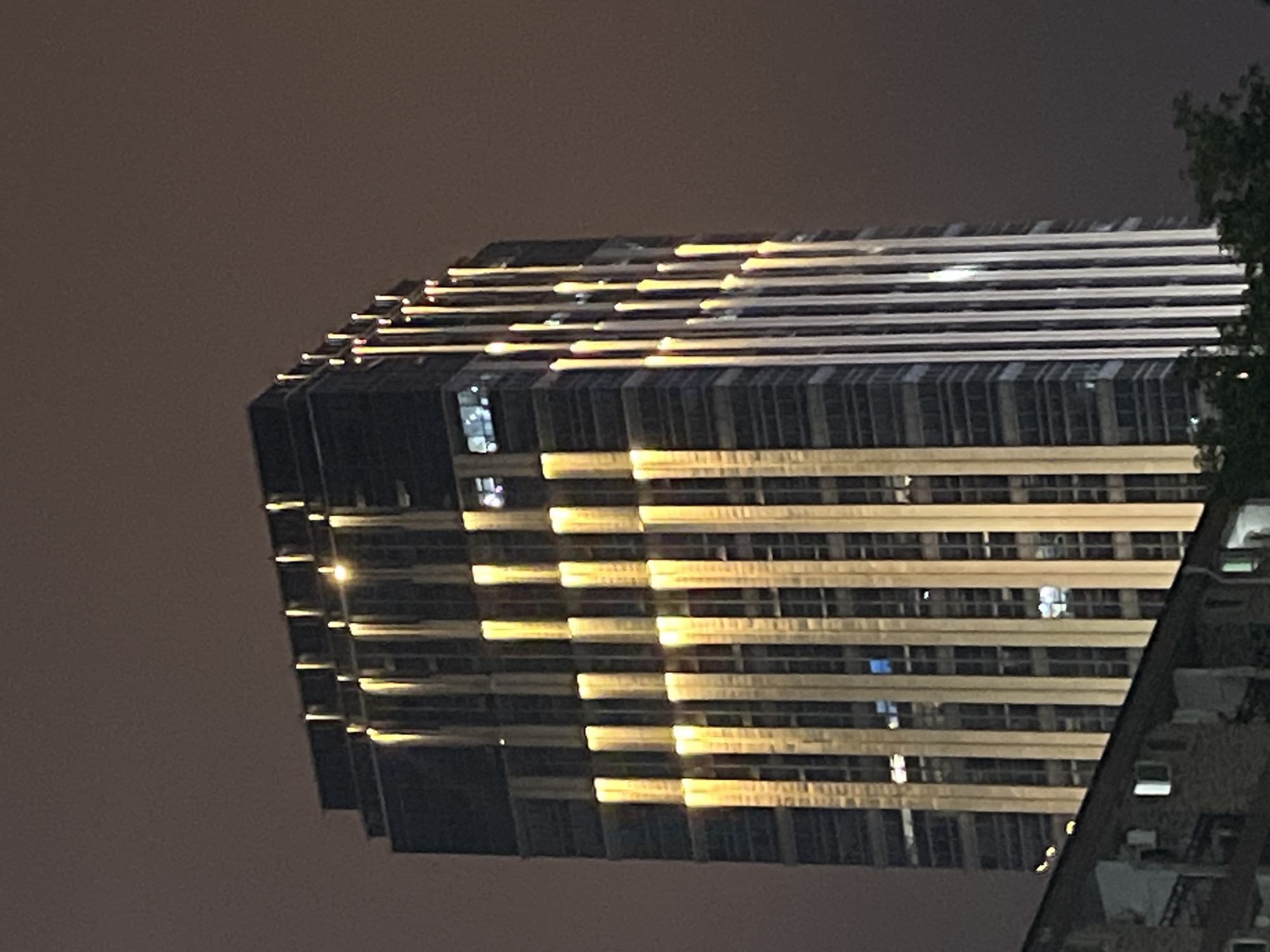 The image shows a tall, modern skyscraper at night, located in the Yuzhong District of China. The skyscraper has a dark facade with numerous windows. Vertical bands of warm yellow light illuminate the exterior of the building, creating a striking visual effect. Some windows within the skyscraper are lit with interior lights, appearing as smaller rectangles of white or blue light. In the foreground, a portion of a lower, older building with brickwork and windows is visible, with some windows also illuminated. Trees are present at the bottom right corner of the image. The sky is dark and appears slightly hazy or overcast, typical of an urban night environment. There are no people clearly visible in the image.