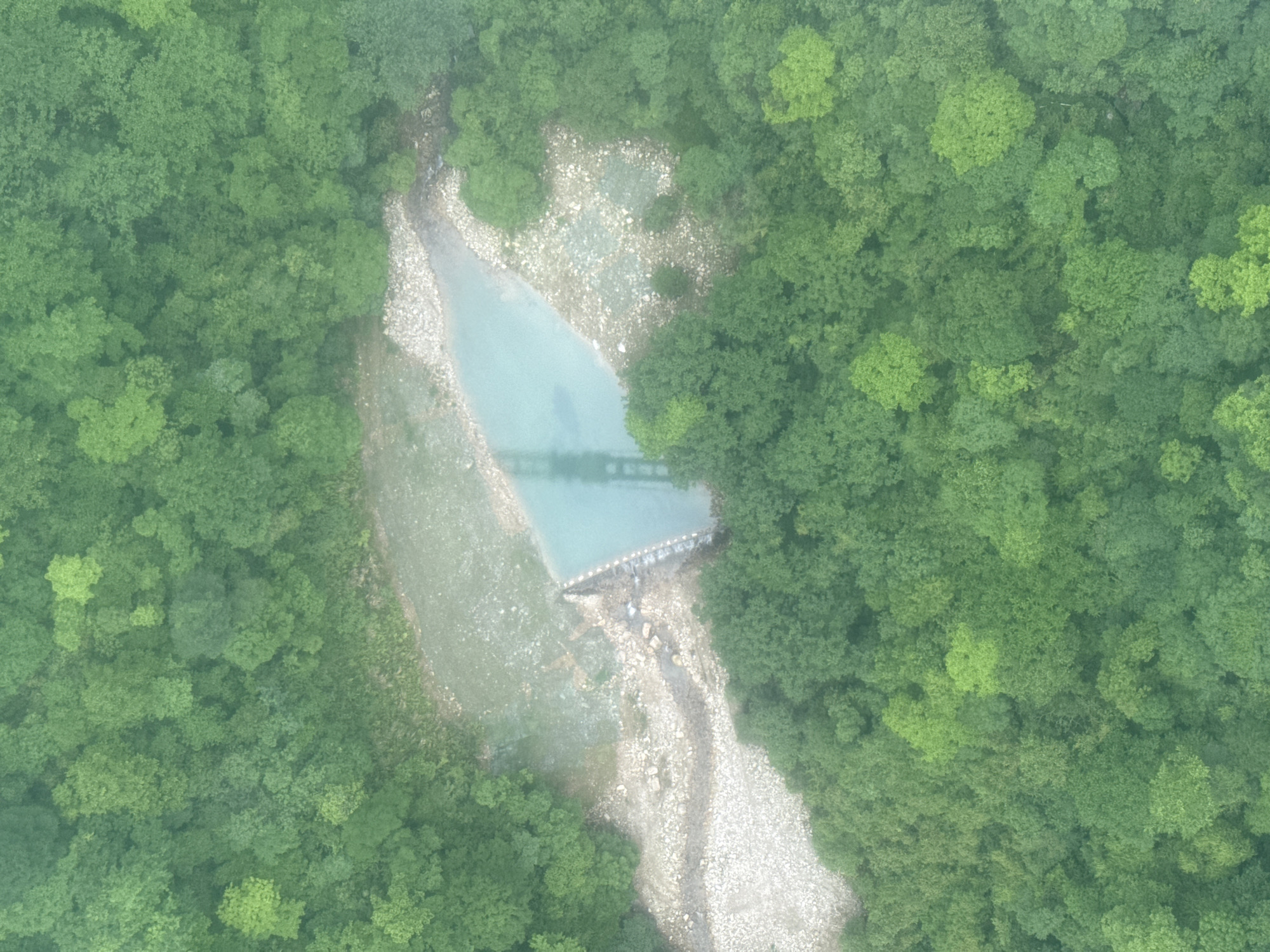 An aerial view shows a narrow river flowing through a dense forest, likely located near the city of Zhangjiajie, China. The river is mostly light blue in color and is surrounded by lush green trees on both sides. A possible dam-like structure is located towards the upper third of the river's flow, causing a pool of water to form just before the structure. There is also a bridge reflecting on the water right before the dam. The riverbed is partially visible and filled with light-colored rocks and pebbles. The photograph exhibits a slightly hazy or foggy atmosphere.