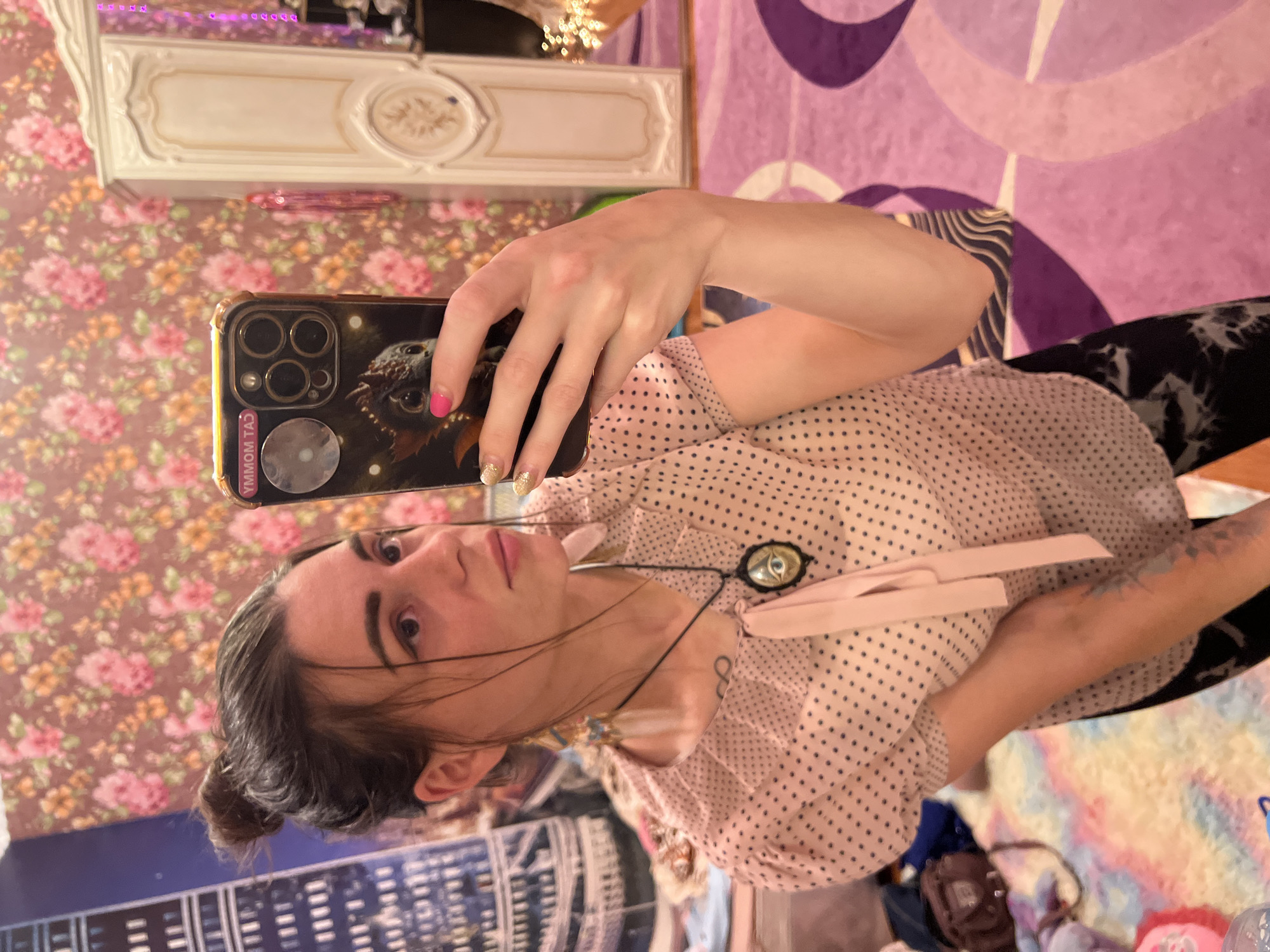 The image is a vertical, wide-angle shot taken indoors, likely in a bedroom. The primary subject is a young woman, captured from the chest up, holding a smartphone. She has dark brown hair pulled back into a bun and is looking directly at the phone's camera, seemingly taking a selfie. Her expression is neutral. She is wearing a light pink blouse with small black polka dots and a ruffled neckline. Around her neck, she wears a pendant with an eye-like design and pink ribbon accents. Her fingernails are painted with bright pink and glittery gold polish.
The background is a busy domestic setting. To the left, there is floral wallpaper with pink and orange flowers on a brown background. Above this, a white ornate piece of furniture, possibly a dresser or shelf, is visible, reflecting some pink light from what appears to be fairy lights or LED strips. To the right, a portion of a purple patterned textile is visible, likely a rug or blanket. Below the woman, a cityscape pattern is visible on what might be a bedspread or wall decoration. Scattered items, including bags and clothing, are visible in the lower portion of the image.
The lighting suggests it is daytime or an interior space with artificial lighting. There are no obvious weather cues. The overall impression is casual and personal.
Visible text on the phone case reads "YMMOM TAJ" in yellow lettering.