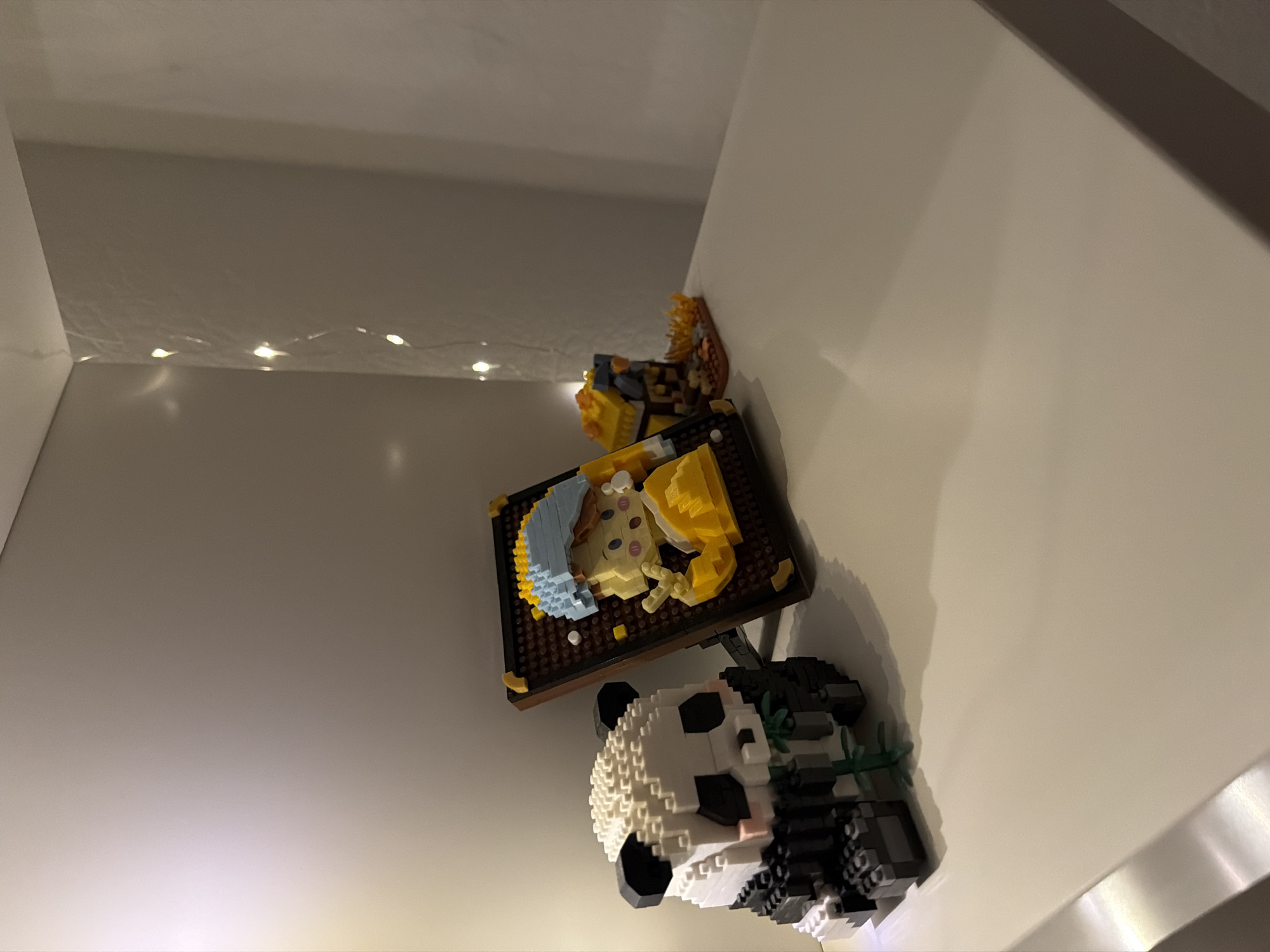 A shelf in an interior space displays several small, pixelated building block figurines. On the left, a panda figurine is positioned to the front, holding green bamboo shoots. To the right of the panda, a framed mosaic portrait of a young person wearing a yellow shirt and a blue cap is propped up. Further to the right, a small, yellow, block-built structure with a fiery orange and brown base sits on the shelf. A string of small, white fairy lights is draped along the wall in the background. The lighting suggests it is evening or a dimly lit room. The scene is devoid of any specific identifiable markers of the city of Kyiv, Ukraine.