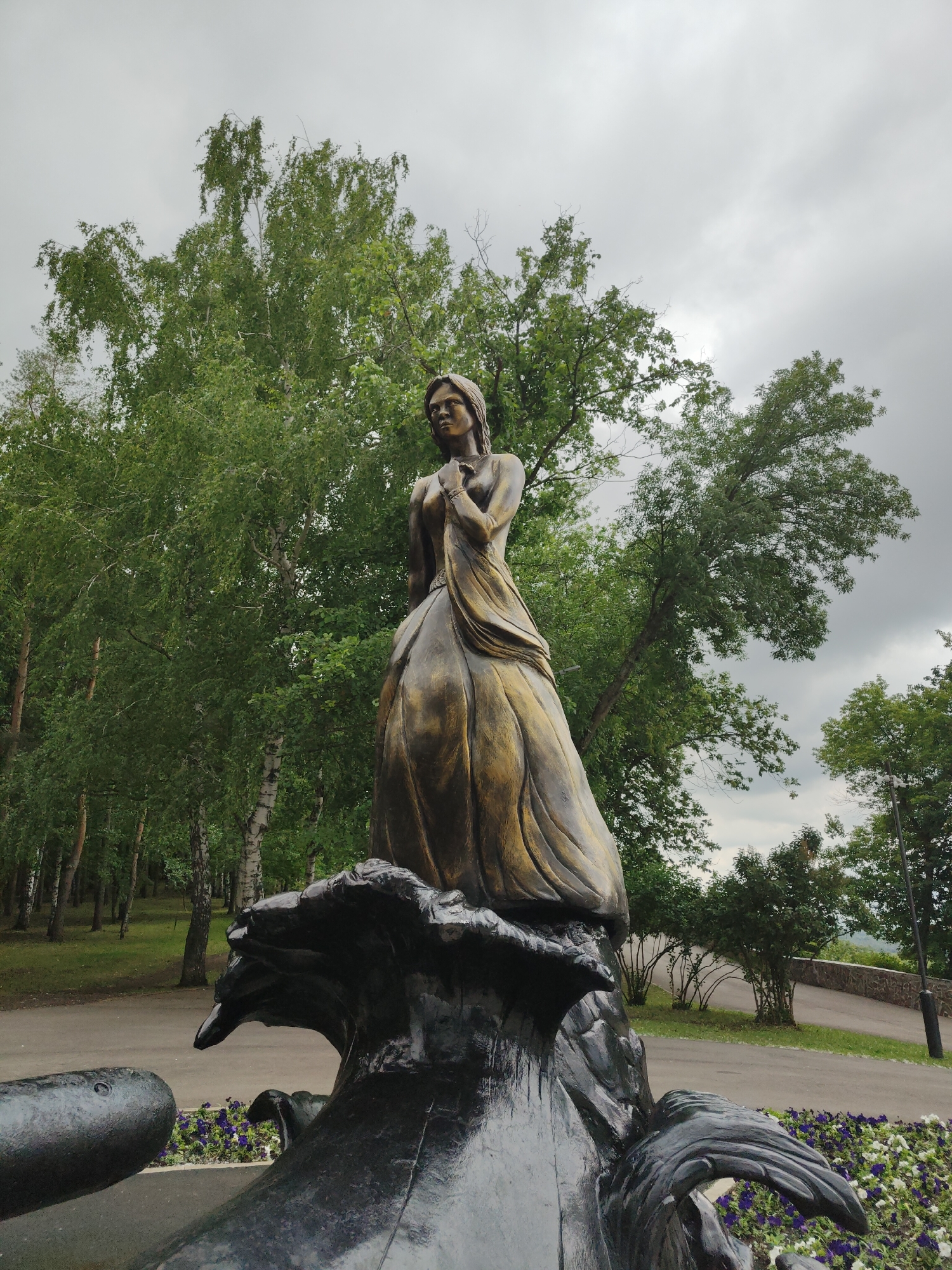 A bronze-colored statue of a woman stands prominently in an outdoor park setting in Unknown, Unknown. The woman in the statue is depicted with long hair, wearing a long, flowing dress, and her hands are clasped near her chest. She appears to be gazing slightly upwards and to her left. The statue is mounted on a dark, sculpted base that features organic, wave-like forms and a prominent, beak-like protrusion on its left side. In the background, a dense array of green deciduous trees, some with white birch trunks, creates a lush canopy. A paved pathway is visible curving through the middle ground, bordered by grass. The sky overhead is overcast and grey. In the foreground, a flowerbed with purple and white blossoms is situated at the base of the statue. The image is captured from a low angle, emphasizing the statue against the natural backdrop.