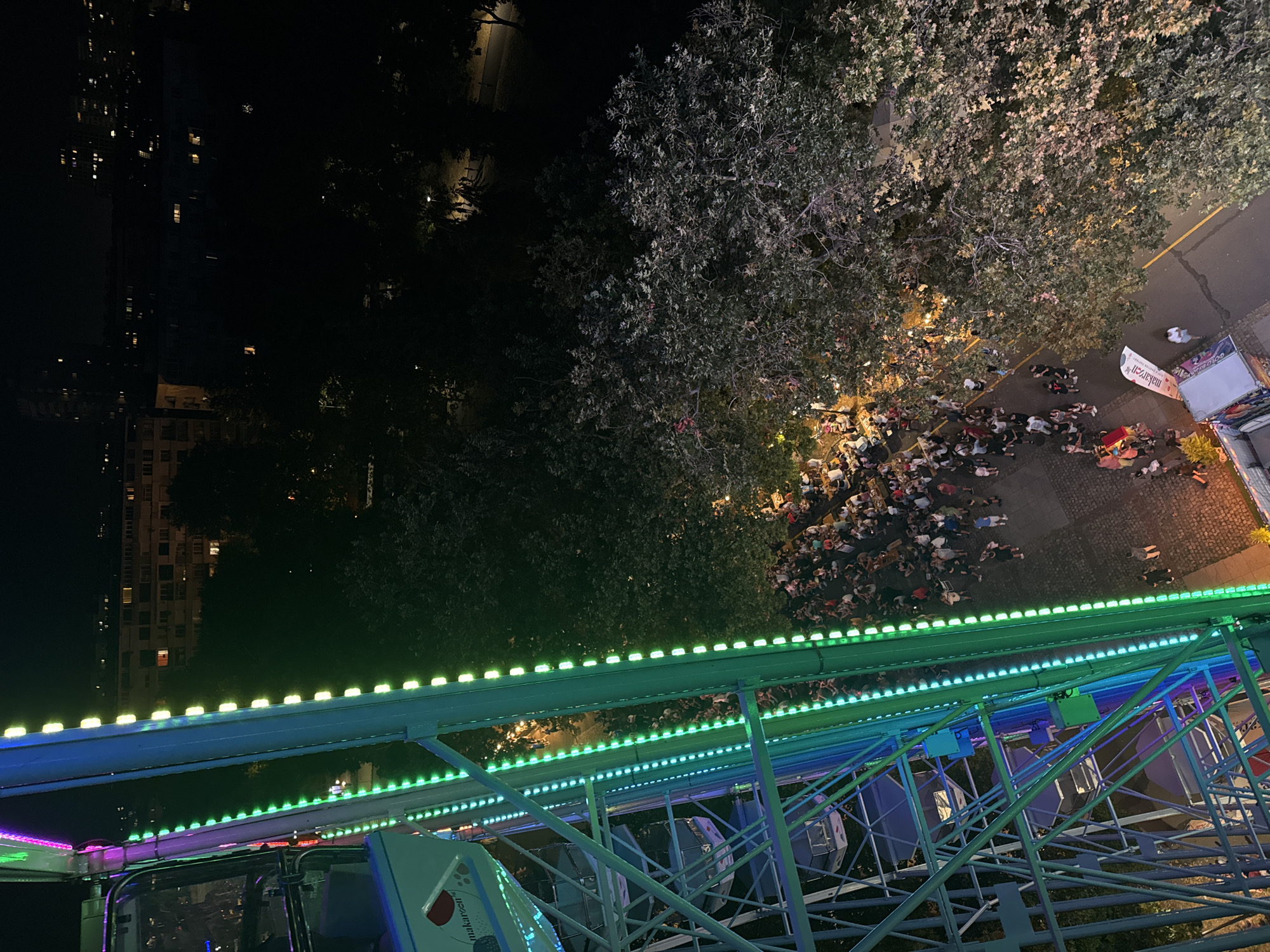 This is a high-angle, long shot image taken at night of a Ferris wheel at an amusement park in Burgas, Bulgaria. The Ferris wheel is brightly lit with green and blue lights, and some of the carriages are visible. Below the Ferris wheel, a crowd of people is gathered, with some walking and others standing in groups. The area is paved, and there are trees with lights illuminating them. In the background, city buildings are visible with some windows lit up. The scene appears to be a lively outdoor event.