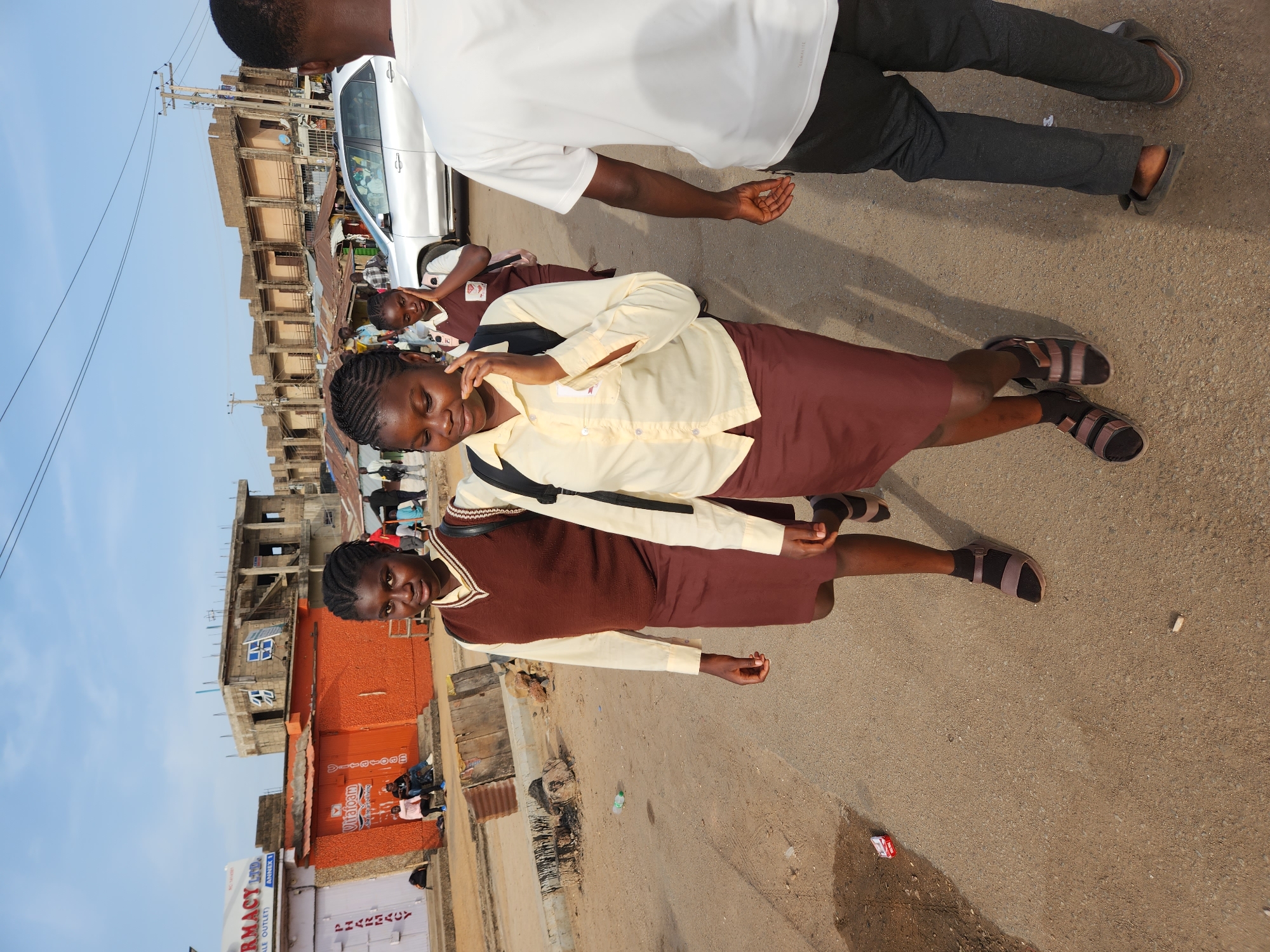 Two schoolgirls in maroon skirts, tan button-down shirts, and brown sweaters walk on a paved street in Kabong, Nigeria. Both are wearing sandals and carrying backpacks. One girl is adjusting her braids. Another individual in grey pants and a white shirt partially obstructs the view of the first girl. In the background are buildings, a white car, and other people. A pharmacy sign is visible on a building. The street appears to be a mix of pavement and dirt.