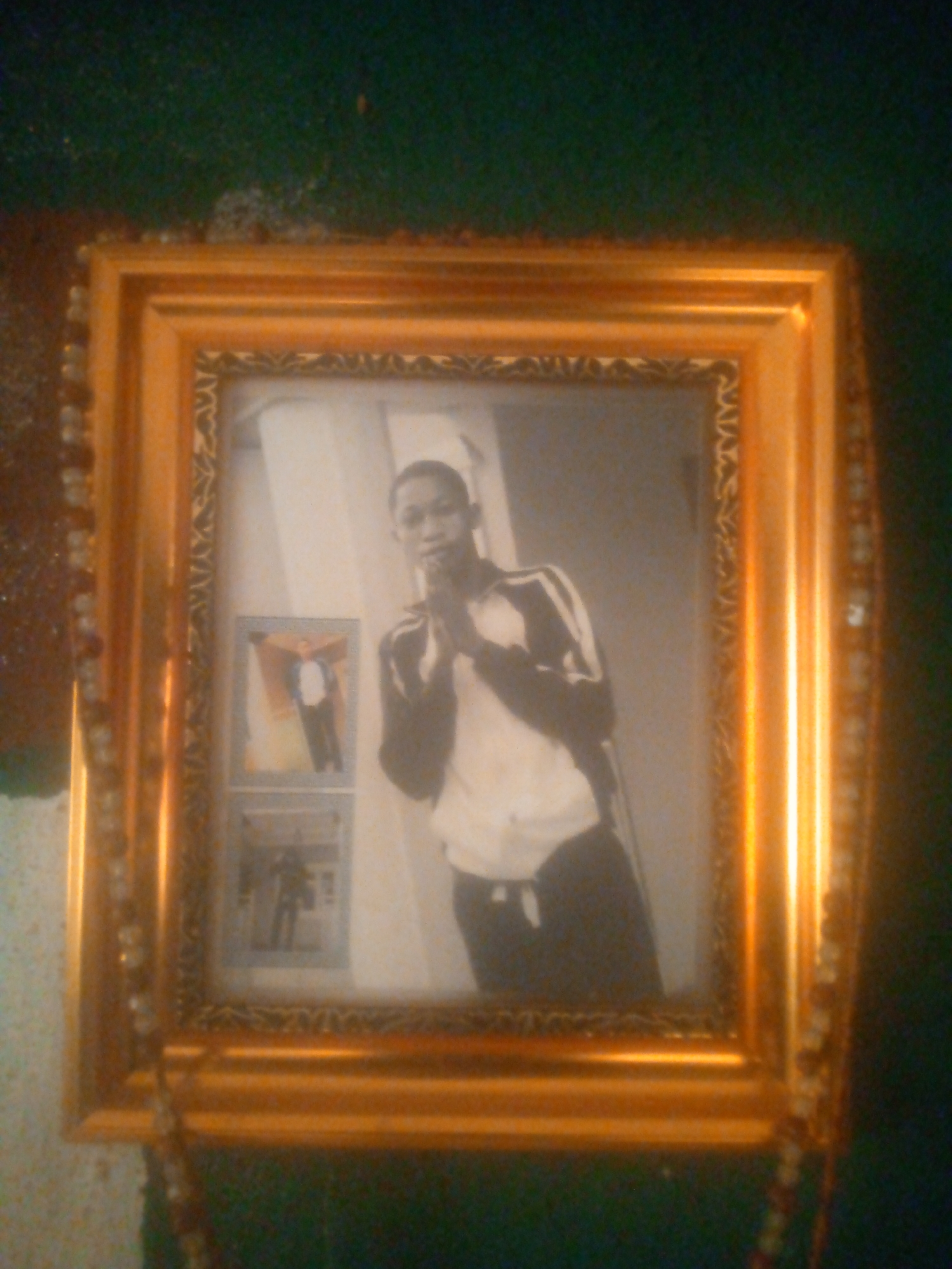 A framed photograph, likely black and white or sepia-toned, is prominently displayed against a dark green wall. The photograph is encased in a large, ornate golden frame with decorative patterns along its inner edge. A string of beads, possibly a rosary or decorative garland, is draped over the left side of the golden frame.
The main photograph features a young, dark-skinned man. He is wearing a light-colored, long-sleeved shirt with dark stripes on the arms, paired with dark trousers. His hands are clasped together in front of his chest or face, conveying a sense of prayer, reflection, or solemnity. In the background of this photograph, two smaller framed pictures are visible on what appears to be a lighter-colored wall. The upper smaller picture clearly depicts a person, possibly a man, dressed in a white shirt and dark pants, standing with arms outstretched. The lower smaller picture is less distinct but also shows a figure.
The overall image is slightly blurry and grainy, suggesting it was taken in low light conditions or with an older camera. The golden frame reflects light, indicating an indoor setting. There is no visible text in the image.