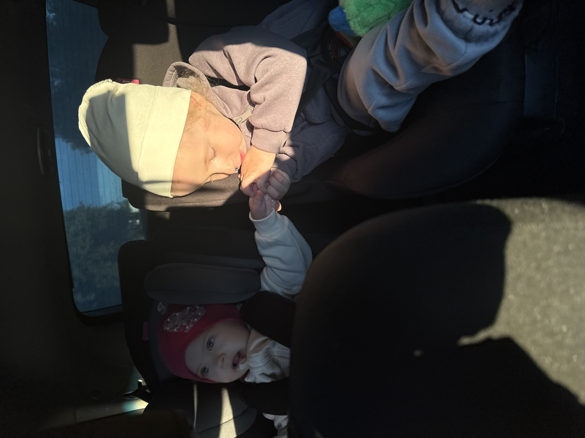 Inside a car in Chaikovsky, Russia, two children are depicted in their car seats, illuminated by strong sunlight streaming through a side window. The image is oriented vertically, showing the back seat.
On the left, an older child, likely a toddler or preschooler, is sleeping peacefully in a car seat. They are wearing a light grey or lavender hooded jacket and a cream-colored soft band or wrap around their head. Their eyes are closed, and their head rests against the car seat's headrest. Their right hand is gently clasped around the left hand of a younger child.
To the right, an infant or baby is secured in a separate car seat. This younger child wears a pink hat adorned with a sparkly embellishment and a white long-sleeved top. Their face is clearly visible, with blue eyes wide open, looking towards the viewer with a curious, slightly open mouth.
Warm, bright sunlight enters the car from the left, casting sharp shadows across the children and the car interior, indicating it is daytime, possibly morning or late afternoon. Through the car window, blurred greenery, possibly trees or distant foliage, is visible. The scene captures a moment of quiet connection and travel between siblings. No visible text is present in the image.