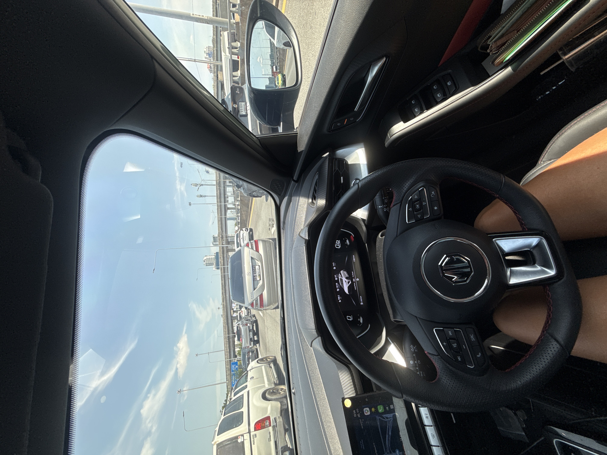 The image shows a first-person perspective from the driver's seat of a car, likely in Bangkok, Thailand, due to the license plate format visible on the car in front and the general urban environment. The driver's hands are on the steering wheel, which has the MG logo in the center. The car is stuck in traffic on an elevated highway, with other vehicles visible ahead and on adjacent lanes. The sky is blue with scattered clouds. The car's dashboard displays navigation information, the current time as 11:14, and other vehicle data. To the right, a side mirror reflects a yellow car and another vehicle on the highway. The interior of the car features black upholstery with red stitching on the steering wheel and seats. A wallet and other items are visible in the passenger door pocket.