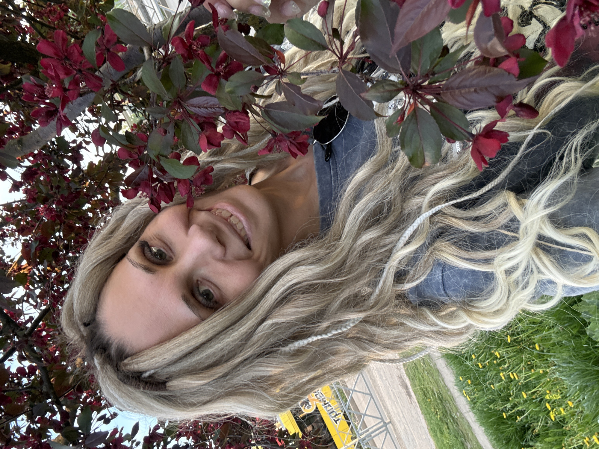 A woman with long, wavy blonde hair with braids is smiling at the camera. She is wearing a dark-colored top and is standing beneath a tree with dark red leaves and flowers. The background shows green grass with yellow flowers, a paved path, and metal structures, suggesting an outdoor area in Rusanovo, Russia.