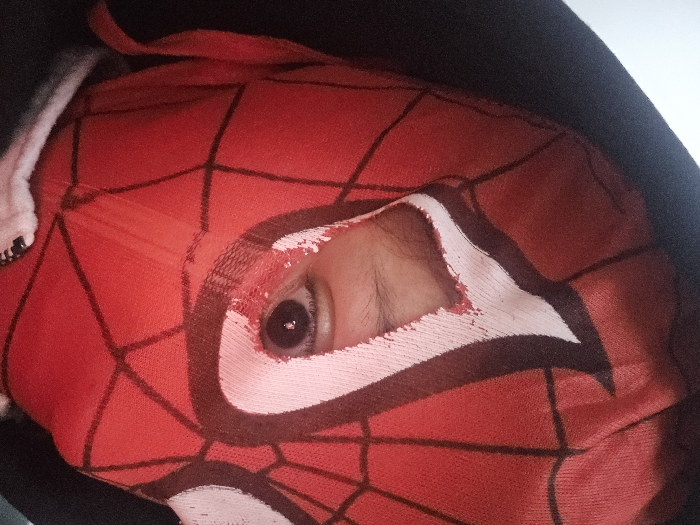 A close-up shows a person wearing a red Spider-Man mask with black web designs. Only one eye is visible through a hole in the mask, peering out. The mask stretches over the face, and the person is wearing a black hooded jacket. The hood is up, framing the mask. Part of a white garment peeks out near the zipper of the jacket. The lighting is soft, highlighting the texture of the mask.