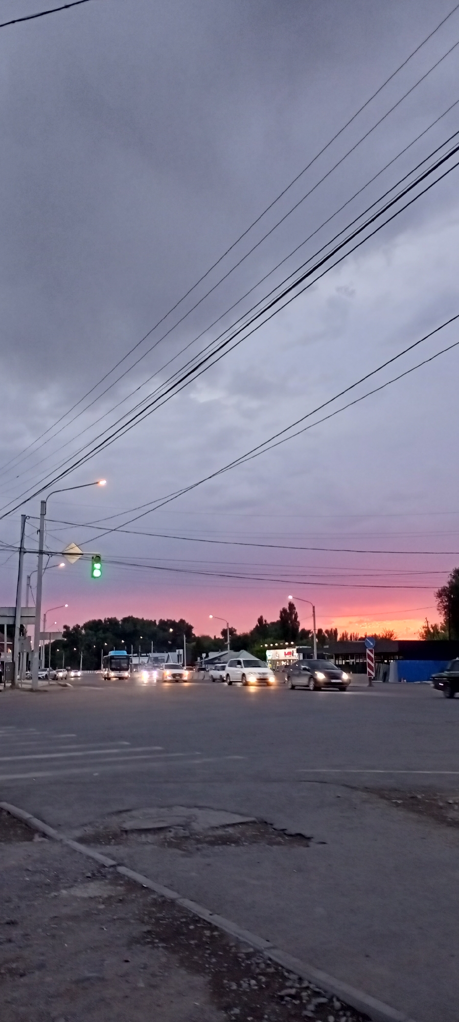 A street intersection in Sokuluk, Kyrgyzstan, is captured during dusk. The sky displays a gradient of colors, transitioning from a pale lavender at the top to a vibrant pink and orange near the horizon, indicative of a sunset. Power lines crisscross the upper portion of the frame.
In the foreground, the asphalt road shows signs of wear, with a prominent pothole and a raised curb. A pedestrian crossing is visible. Traffic lights are present, with the green light illuminated on one.
Several vehicles are on the road. A blue bus is in motion, as are multiple cars. Some vehicles have their headlights on, illuminating the road ahead. Buildings and trees form the backdrop, suggesting an urban or semi-urban environment. The lighting conditions are dim, typical of twilight. No people are clearly discernible in the image.