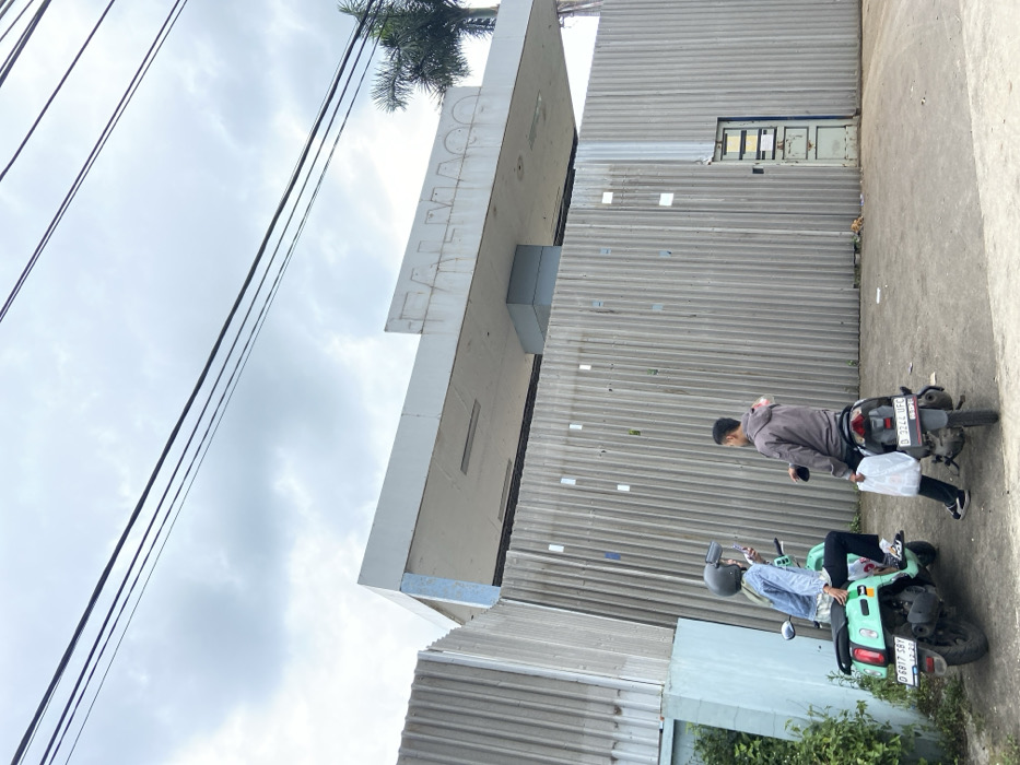Two people are standing in front of a corrugated metal fence. The fence is in front of a building with a sign that says "FALMAQ". One person, a woman wearing a hijab and a denim jacket, is standing on a teal moped. The other person is a man wearing a grey jacket and is standing next to a black moped. Both mopeds are parked. Both people appear to be interacting. The sky is cloudy. There are power lines overhead. There are two palm trees in the background. This scene is located in Ngamprah, Indonesia, at coordinates -6.861, 107.5.