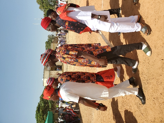 The image displays an outdoor scene in Bar Gada, Nigeria, under a clear blue sky and bright daylight. The foreground features five men walking from left to right across a dry, light brown dirt ground. The leftmost man, partially visible, wears a white tunic, white trousers, black dress shoes, and a red patterned cap, holding a dark object in his left hand. The second man wears a black short-sleeved shirt with orange and red patterns and a red and white striped cap. The third man, centrally located, wears a similar black and orange patterned short-sleeved shirt, dark trousers, a red and white striped cap, white socks, and olive green sandals, holding a dark object in his right hand. The fourth man wears a white tunic and trousers, a red cap, dark sandals, and has a black and red patterned fabric draped over his left shoulder, carrying a light-colored staff in his right hand. The fifth man, partially visible, wears a white tunic. In the left mid-ground, multiple individuals are seated on white plastic chairs under a green canopy. Behind them, a light-colored, multi-story building with windows is visible. Trees and additional individuals are present in the background, to the left and right of the building. Two thin poles are observable in the far right background.