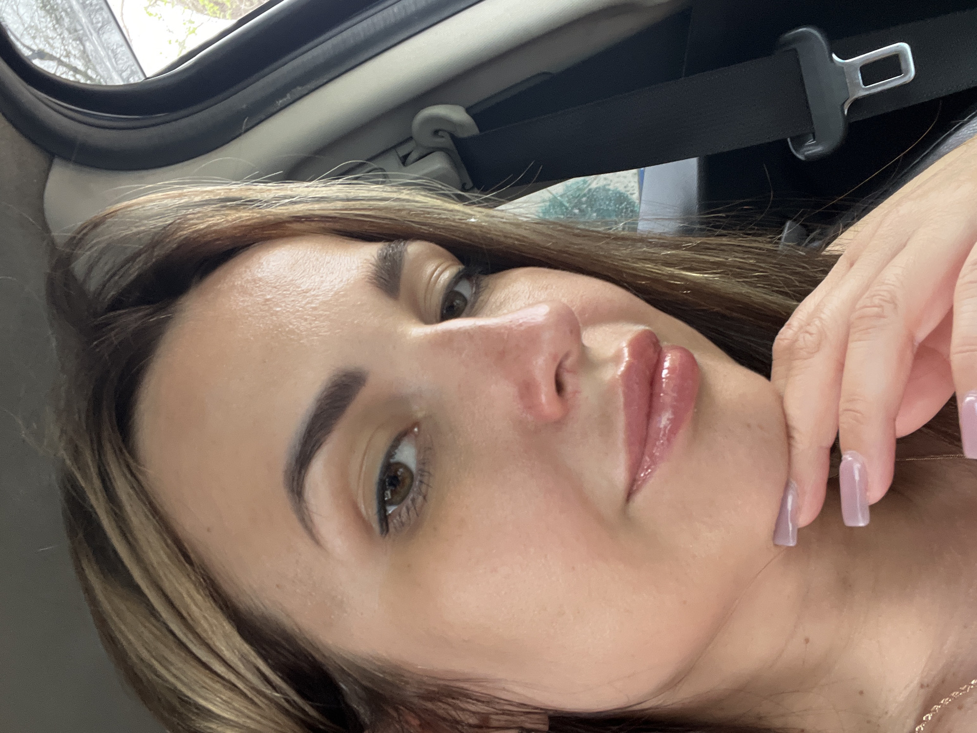 A close-up, waist-up, indoor selfie shows a woman with light brown hair and brown eyes looking slightly to the left. She is wearing a light pink lip gloss. Her right hand is resting on her chin, and her fingernails are painted a light purple. She is wearing a necklace. A black seatbelt is visible behind her on the right side of the frame. The background suggests she is in a vehicle, with a car door and a blurred outdoor scene visible through the window.