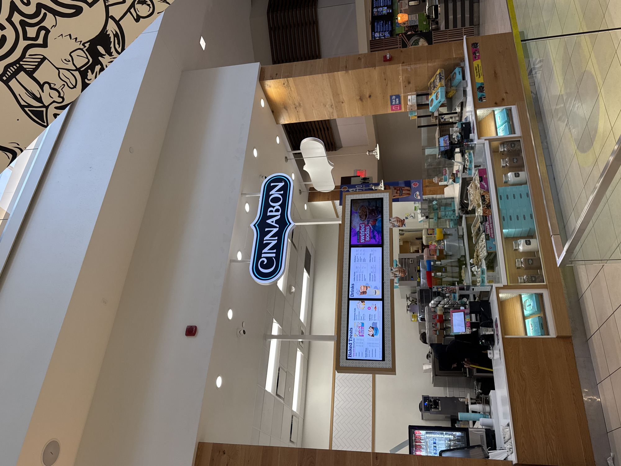 The image depicts a Cinnabon store situated within a brightly lit, spacious indoor complex, likely a mall. The store is characterized by its prominent, vertical "CINNABON" sign, featuring glowing blue and white letters, suspended from the high ceiling.

The store's counter is constructed from light-toned wood, extending across the frame. Display cases along the counter showcase various Cinnabon products, including boxes and jars, many in light blue packaging. A "Pick Up Here" sign is visible on one section of the counter.

Above the service area, two large digital menu boards are clearly displayed. The left board lists "Baked Treats," detailing items such as "Classic Rolls," "MiniBons," "Cinnabon Bites," "Center of the Roll," and "Cinnasweeties," with associated prices. The right board presents "Drinks," including options like "MochaLatta Chill," "Caramel PecanBon," "Cold Brew Iced Coffee," "Lemonade," and "Water." A smaller screen above these advertises "Try Our NEW FROSTED SODAS" and "Jumbo Swirled Sodas."

Behind the counter, an employee in dark attire is actively working, with various kitchen equipment, dispensers, and supplies visible.

The surrounding environment features high white ceilings dotted with numerous recessed lights, contributing to the bright atmosphere. An artistic black-and-white mural, depicting abstract, cartoon-like figures, adorns a sloped wall in the upper left corner of the image. The floor is tiled in a light pattern. In the background, hints of other commercial establishments and architectural elements, such as wooden slats, suggest a bustling commercial setting. The time of day appears to be daytime, given the bright interior lighting.