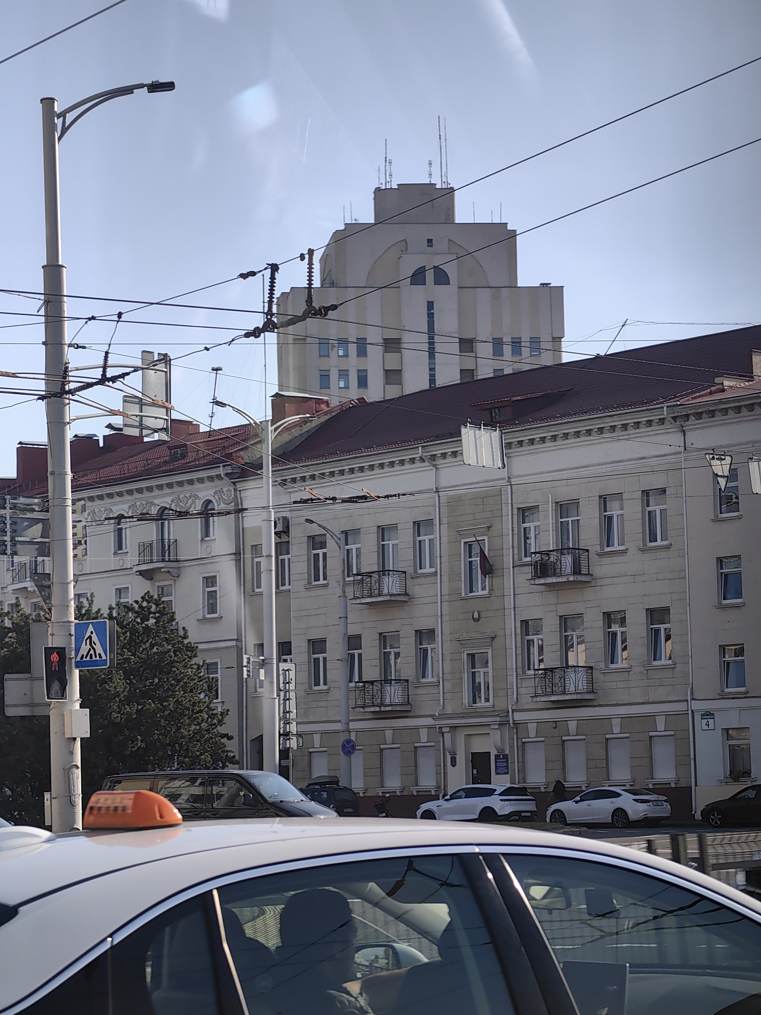 The image captures a street scene in Minsk, Belarus. In the foreground, the roof of a white car is visible, with an orange taxi sign on top. Behind the car, a street with overhead power lines and a traffic light are present. A pedestrian crossing sign is also visible. Several buildings line the street, featuring architectural details such as balconies and ornate facades. The buildings are of varying heights, with a larger, stepped building in the background. Several cars are parked along the street, including white sedans and an SUV. The sky is clear and blue. The image appears to be taken from a moving vehicle, possibly a taxi, due to the perspective and the reflection on the car's surface.