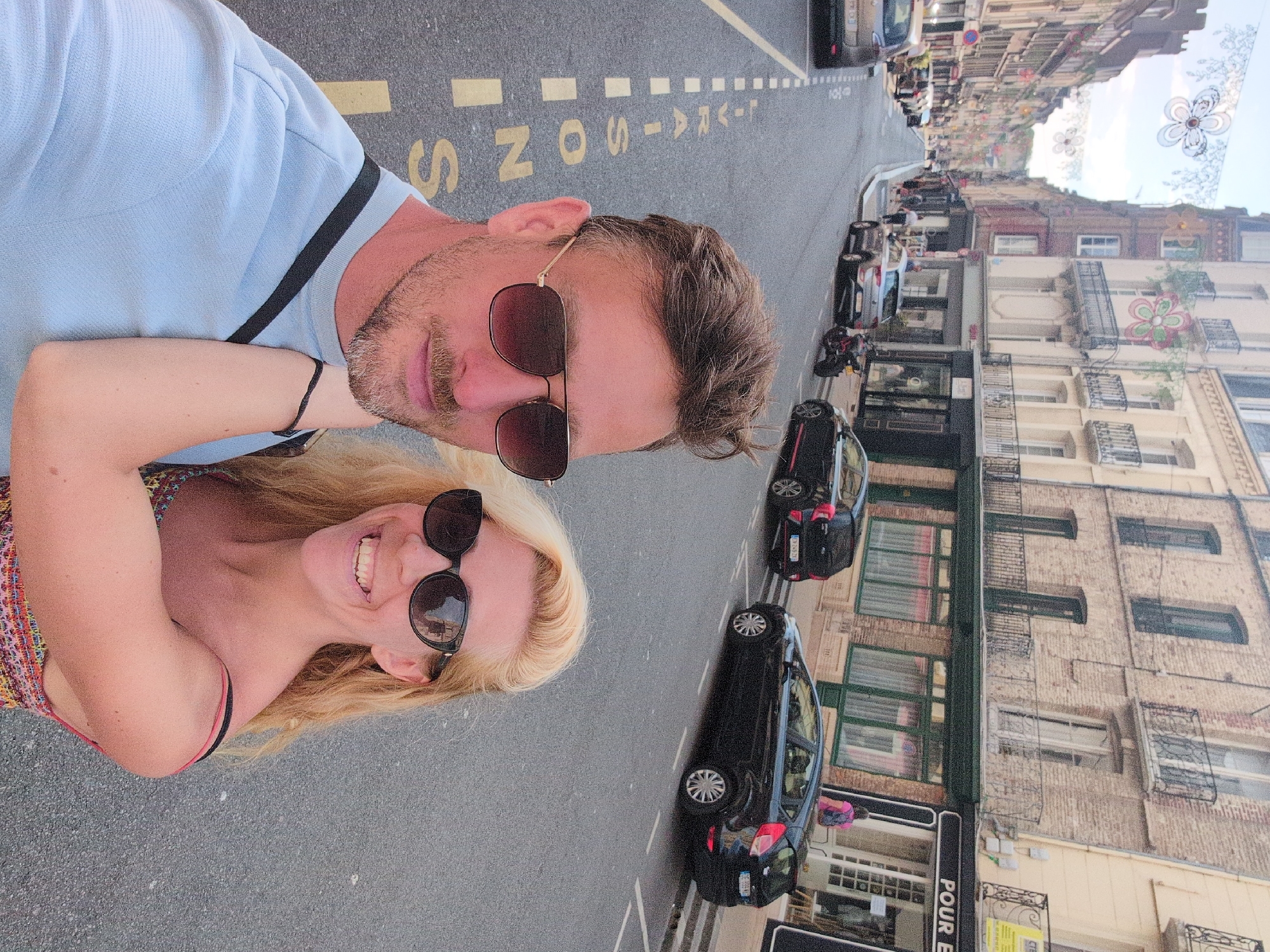 A selfie is taken by a couple standing on a street in Dieppe, France, possibly during a festival. The man, on the left, wears sunglasses and a light blue t-shirt, with his arm around the woman. The woman, on the right, also wears sunglasses and a colorful, strappy top, smiling at the camera. Behind them, a narrow street is lined with traditional French buildings with balconies and awnings. Several parked cars and a motorcycle are visible on the street, and decorative flower garlands hang overhead, suggesting a festive occasion. The road has white dashed lines marking parking spaces and yellow painted text, which reads "L-AVAISONS" and "Z N S".