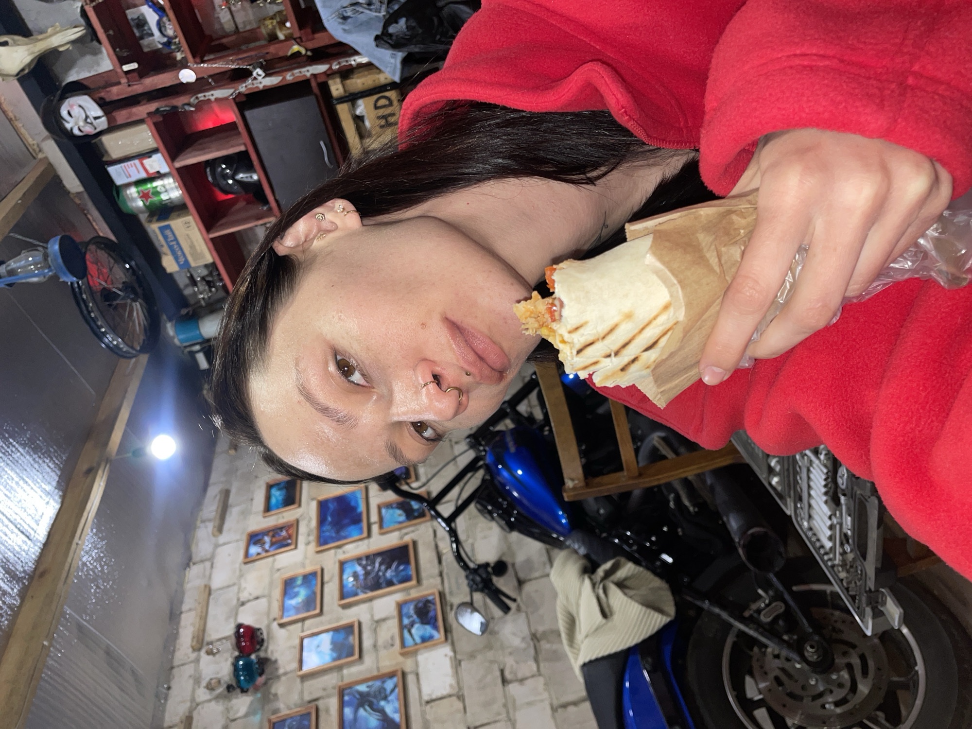 A person in a red garment is eating a wrap. The person has a fair complexion, brown eyes, and dark hair. They have multiple piercings in their ears and nose. The wrap is partially eaten and wrapped in paper.
The background appears to be a garage or workshop setting. There's a blue motorcycle parked behind the person. The walls are made of light-colored brick and decorated with framed pictures. There is a shelf with various items, including boxes and containers, on the upper left of the frame.
Given the query, the location is Oktyabr'skii, Russia. No further location details are discernable from the image.