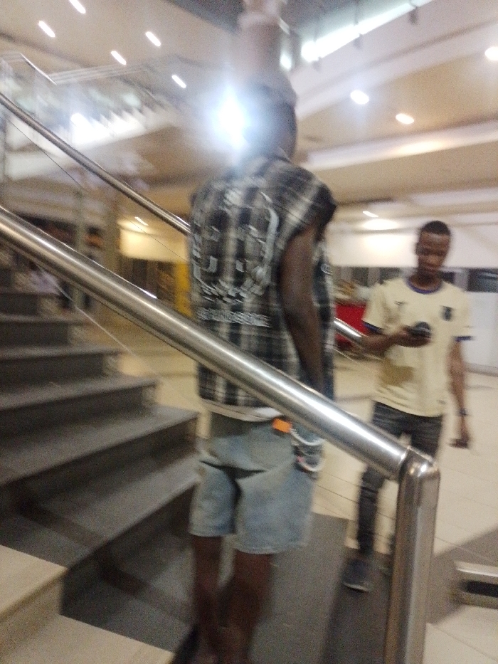 The image depicts an indoor, multi-level architectural space within Gandu Albasa, Nigeria. In the foreground, a dark-skinned male is viewed from the rear, positioned beside a set of dark-colored stairs and a metallic handrail. He wears a sleeveless black and white plaid shirt with a graphic design on the back and light blue denim shorts; an orange object is visible on his right hip. A bright light source creates a glare over his upper back and head area. In the mid-ground, a second male subject, lighter in complexion, stands facing right while looking down at a dark-colored smartphone held in his hands. He wears a light-colored short-sleeved t-shirt with a blue collar and a dark emblem on the chest, paired with dark pants. The environment features a light-colored ceiling with multiple recessed lights. Blurred architectural elements, including additional stairways or an escalator, are discernible in the background.