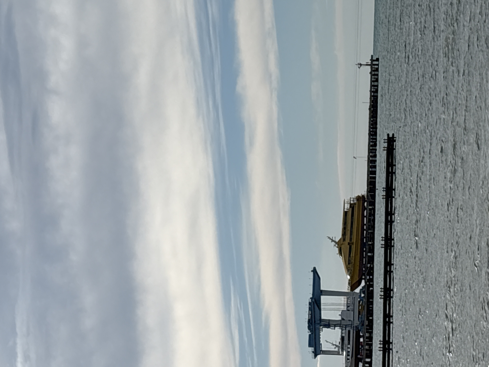 The image shows a view from Anapa, Russia, of a pier extending into the sea. A yellow passenger ferry is docked on the pier. A crane or loading structure is situated at the base of the pier. The sky is mostly covered with streaky clouds. The sea appears calm with some slight ripples visible.