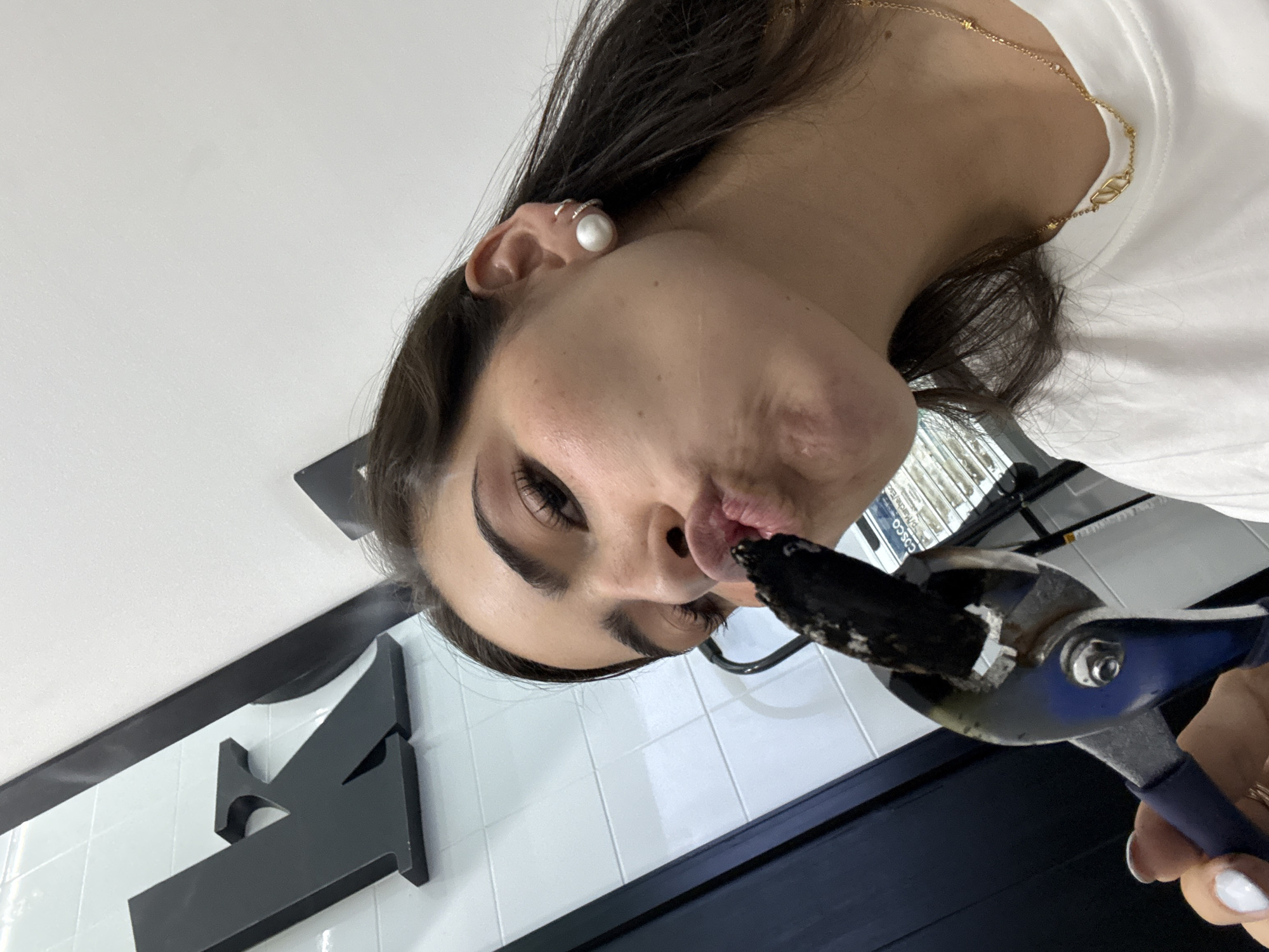 A woman holds a piece of burning charcoal with blue-handled pliers in what appears to be an indoor space with white tiled walls and a partial black sign. The woman has dark hair, wears a pearl earring, and a gold chain necklace. Smoke rises from the charcoal. The location is San Bernardino Tlaxcalancingo, Mexico.