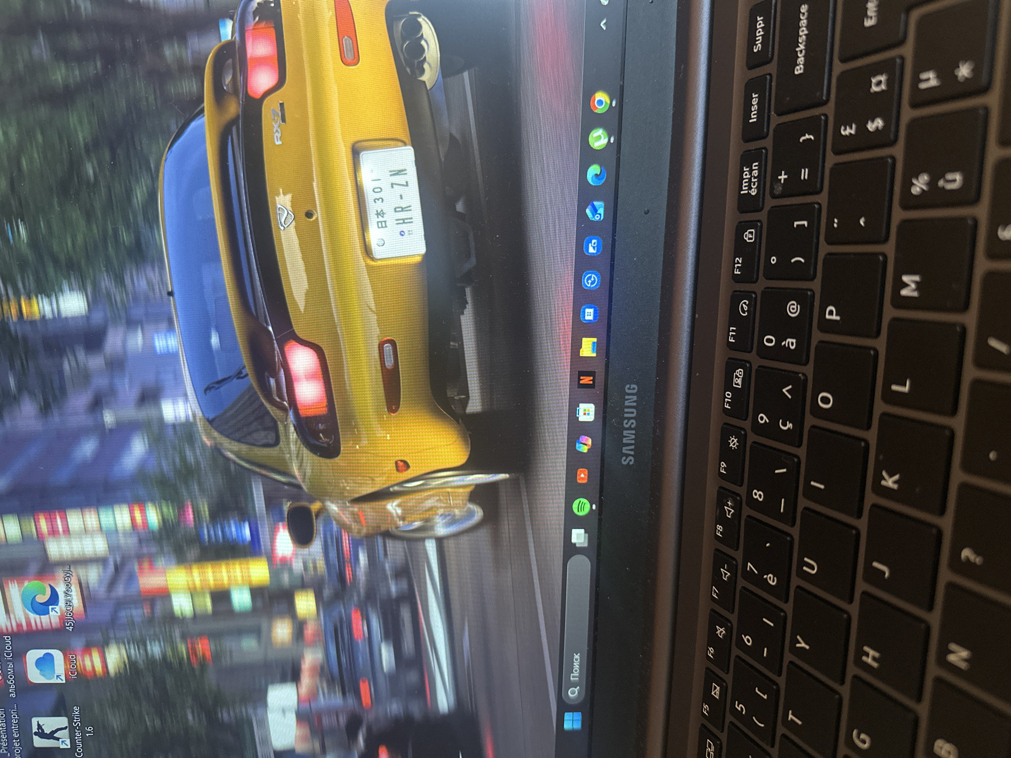 The image shows a laptop with a desktop wallpaper featuring a yellow Mazda RX-7 driving in a city at night. The wallpaper depicts a vibrant urban scene with blurred lights from buildings and other vehicles, creating a sense of motion. The car's taillights are illuminated, and its license plate is partially visible.
On the laptop screen, a taskbar is visible at the bottom. It includes a search bar with the Russian word "Поиск" (Poisk), meaning "Search," and several application icons such as Spotify, YouTube, and Microsoft Edge. The laptop itself is a Samsung model, as indicated by the logo on the bezel above the keyboard. The keyboard is a QWERTY layout with some keys showing French characters.
The image does not contain any information that would place the scene in the city of Anduze, France.