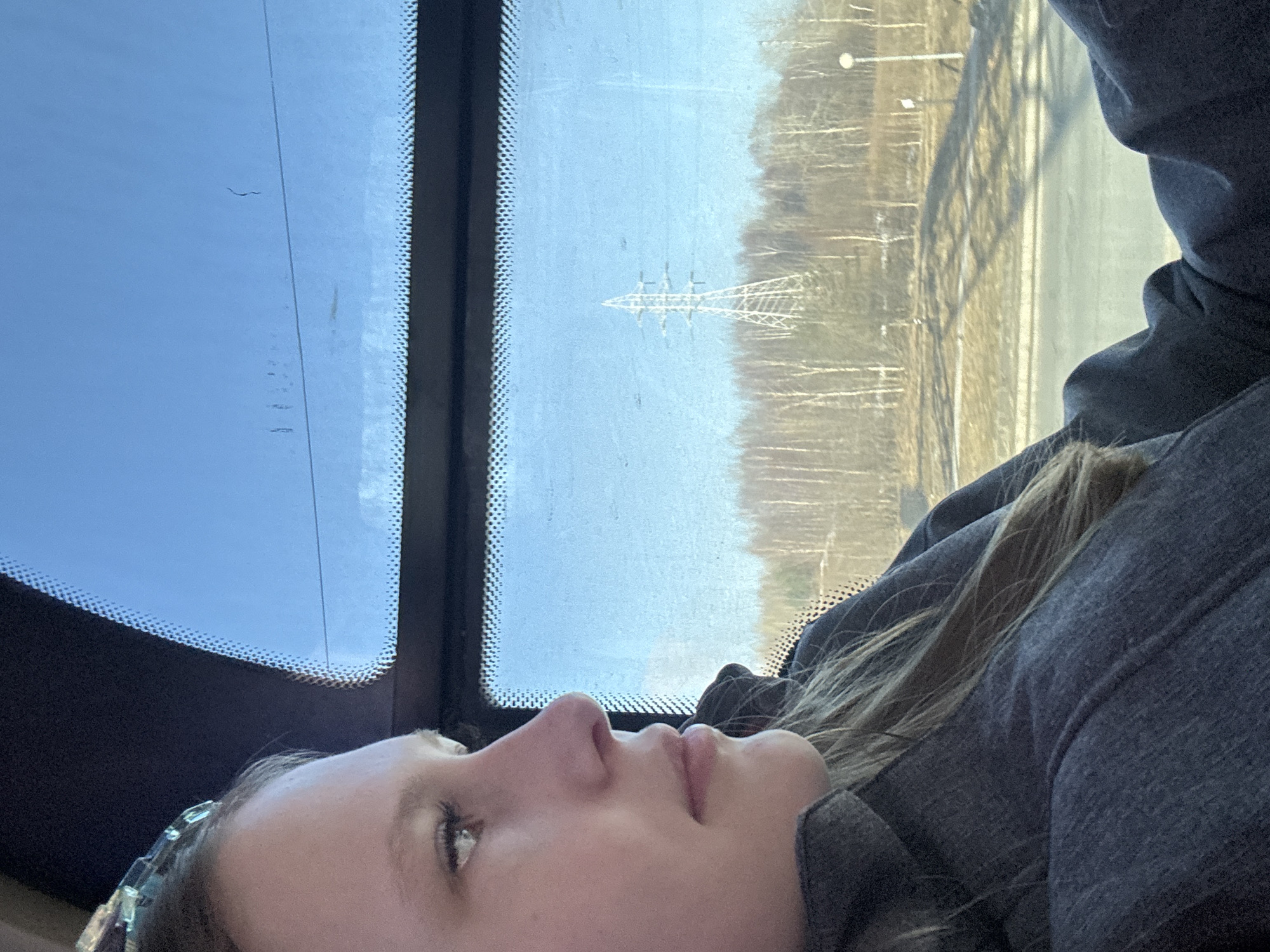 A person with fair skin and light brown hair is visible in the lower portion of the frame, partially obscured by a gray hooded garment. The person appears to be looking upwards towards a clear sky, which is visible through a vehicle windshield. In the background beyond the windshield, there is an electrical transmission tower visible against a backdrop of sparse trees and possibly a road or bridge structure. The photo seems to be taken from inside a moving vehicle. Given the prompt's request to identify the location as Usolye, Russia, the photo likely depicts an individual within a vehicle in that area, observing the surrounding landscape which includes the transmission tower.