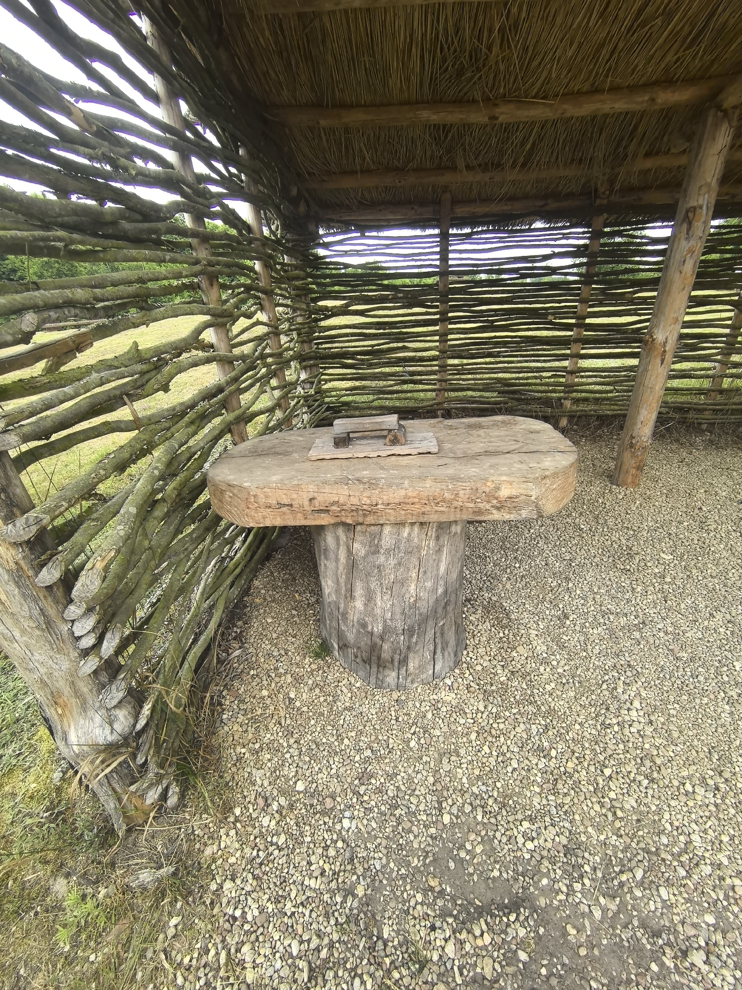 The image depicts an outdoor scene within a rustic structure that appears to be made from natural materials. The structure has a thatched roof supported by wooden beams, and its walls are constructed from horizontally woven branches. In the center of the structure, a rough-hewn wooden table stands on a thick, log-like base. The table top is wide and flat, with a visible seam or crack. On the table's surface, there are two small, rectangular wooden objects. The ground within the structure is covered with small, light-colored gravel. To the left of the structure, a grassy area is visible, with some dry grass and foliage around the base of the structure. The background shows a green, grassy field with a line of trees or vegetation in the distance, and the sky is overcast. The scene suggests an outdoor seating area or a primitive workstation, possibly in a rural or historical setting. The location is specified as Камянюцкі сельскі Савет, Belarus.