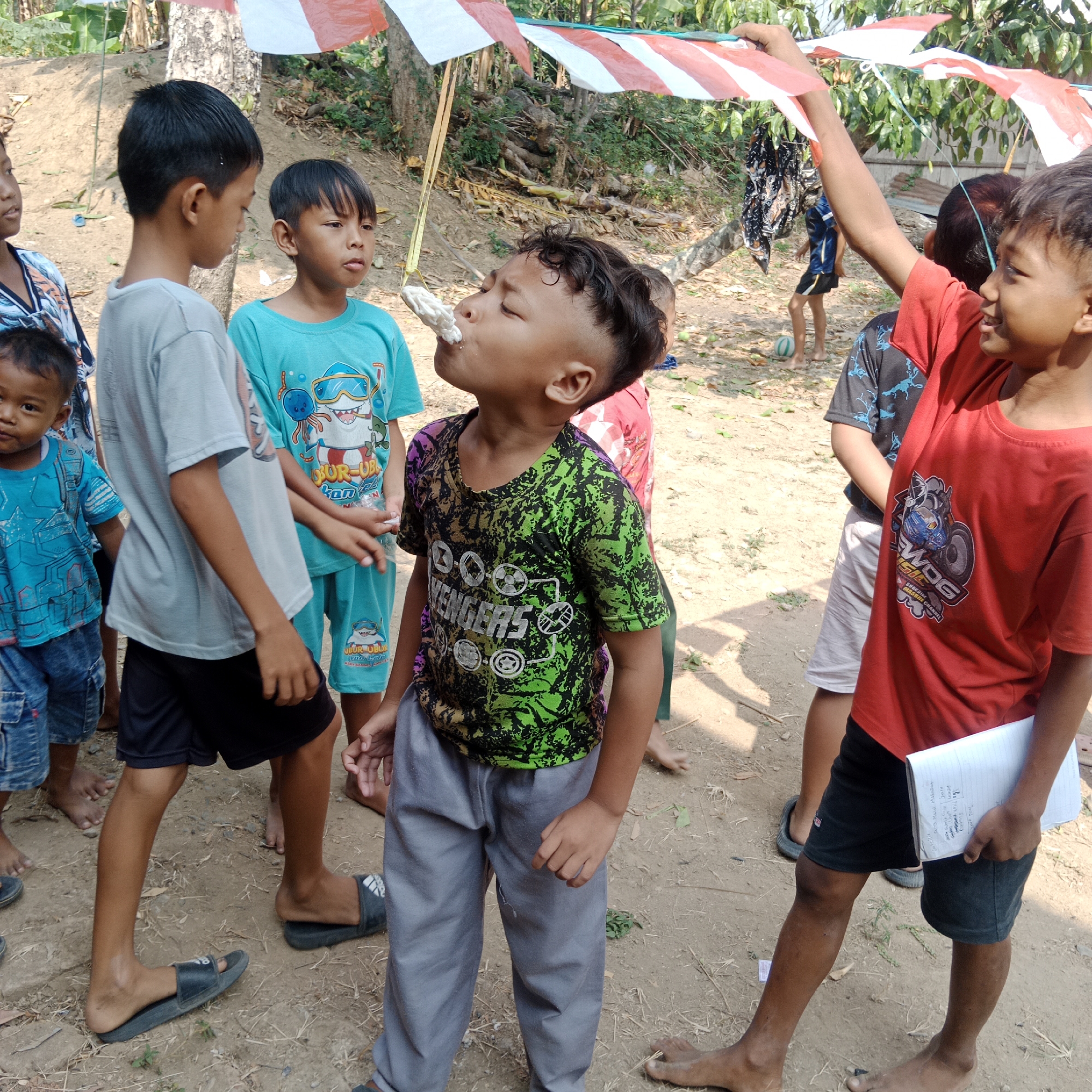 In Tegowanu Kulon, Indonesia, a group of boys is participating in a festive outdoor activity. In the foreground, a boy in a green and black Avengers t-shirt is attempting to eat a white, irregular-shaped food item suspended from a yellow string. He is looking upwards with his eyes closed, focused on the task. To his left, another boy in a turquoise t-shirt with a shark graphic is observing. A third boy in a grey t-shirt and black shorts stands to the far left, also watching the event. Behind them, a red t-shirt adorned with a graphic of a motorcycle and the word "WOG" is visible on another boy. The ground is dry and dusty, with scattered leaves and dry grass. Overhead, a makeshift canopy of red and white striped fabric is set up, possibly for a celebration or event. Various trees and foliage form the backdrop. The overall scene suggests a cultural celebration or game taking place in a rural Indonesian setting.