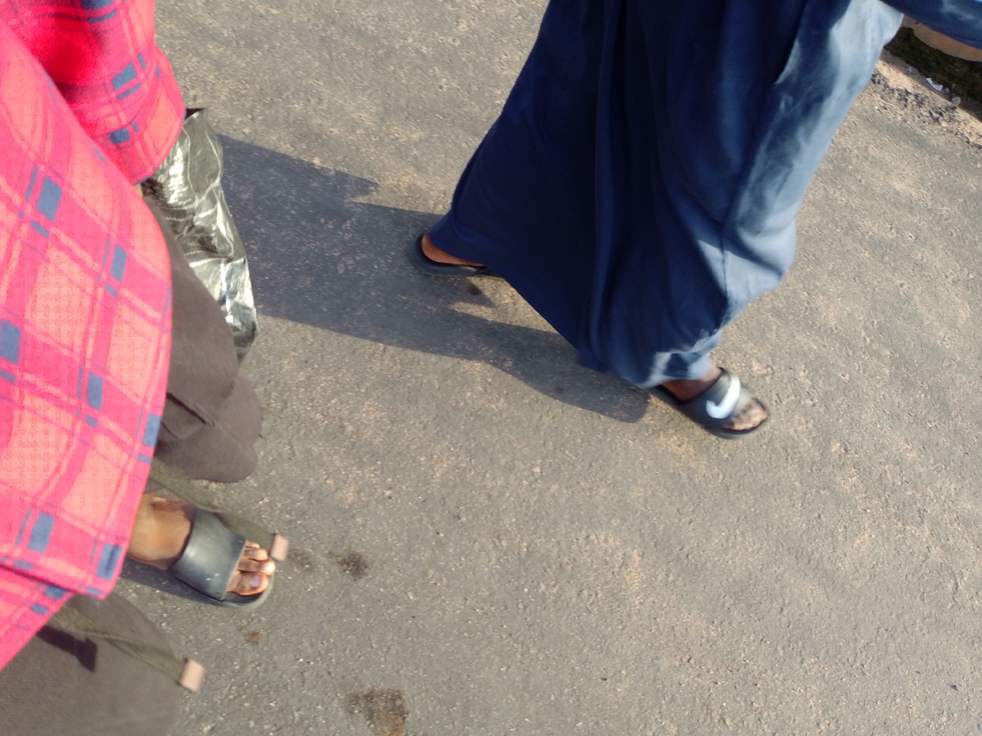 The image shows two people's legs and feet standing on a paved surface. The person on the left is wearing a red and blue plaid garment and black slip-on sandals, with their left foot visible. To the right of this individual, a second person is wearing a long, dark blue garment and black slip-on sandals with a white Nike logo, with their right foot visible. A dark, crinkled plastic bag is positioned between the two individuals, partially obscured by the garment of the person on the left. The ground appears to be asphalt or concrete, with some dirt and debris present. The scene is outdoors, under bright sunlight, casting shadows. Without additional information, the precise location within Jalingo, Nigeria, cannot be determined, but it appears to be a street or walkway.