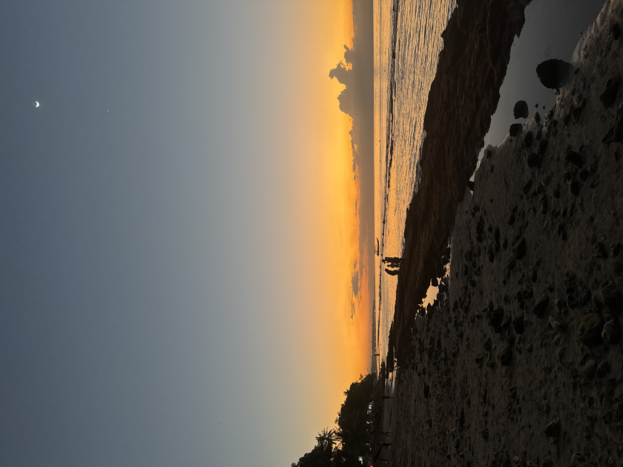 The image depicts a serene beach scene at dusk in Nusa Penida, Indonesia. The sky is a gradient of deep blue to vibrant orange, with a crescent moon and a small, bright celestial body visible in the upper left quadrant of the sky. The ocean, reflecting the warm hues of the sunset, meets a rocky shoreline. Silhouettes of a few people are visible along the water's edge and further back on the sandy beach, suggesting a peaceful evening stroll or gathering. Lush palm trees line the edge of the beach in the foreground, their dark forms contrasting with the bright sky and sea. The overall mood is tranquil and contemplative, characteristic of a sunset on a tropical island. There is no visible text in the image.