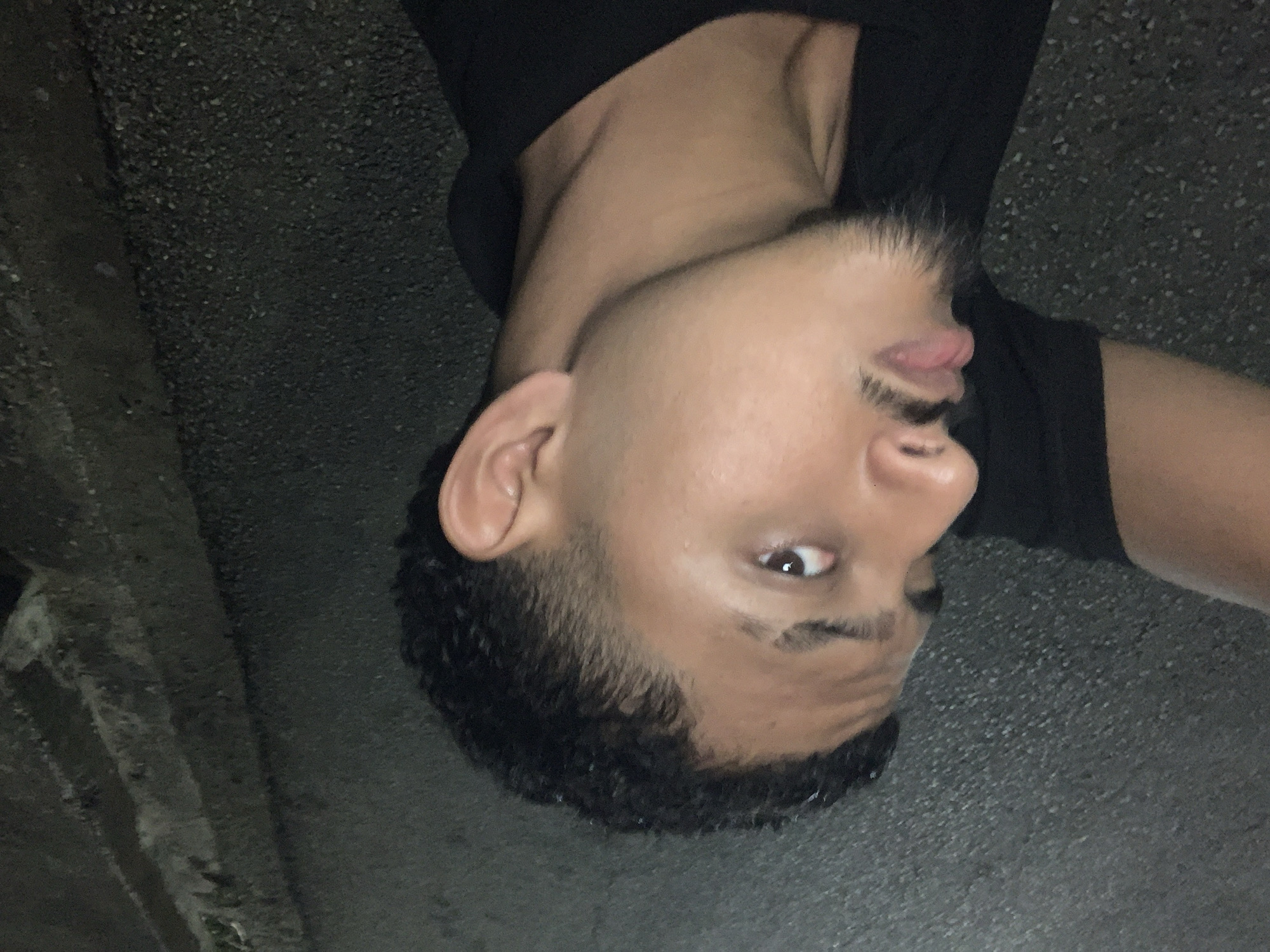 The image shows a close-up, profile view of a young man's face and upper torso, taken outdoors at night, with a dark, granular surface in the background that appears to be asphalt or pavement with some lighter-colored debris or markings. The man has dark, curly hair styled with a fade haircut, prominent eyebrows, a light mustache, and a goatee. His left eye is visible, looking towards the camera. His mouth is slightly open, with his lower lip protruding slightly. He is wearing a black collared shirt. The lighting suggests a low-light environment, likely street lighting or artificial light sources. The background is dark and indistinct, with subtle variations in texture and shadow. The location is identified as Santa Ana, Venezuela.