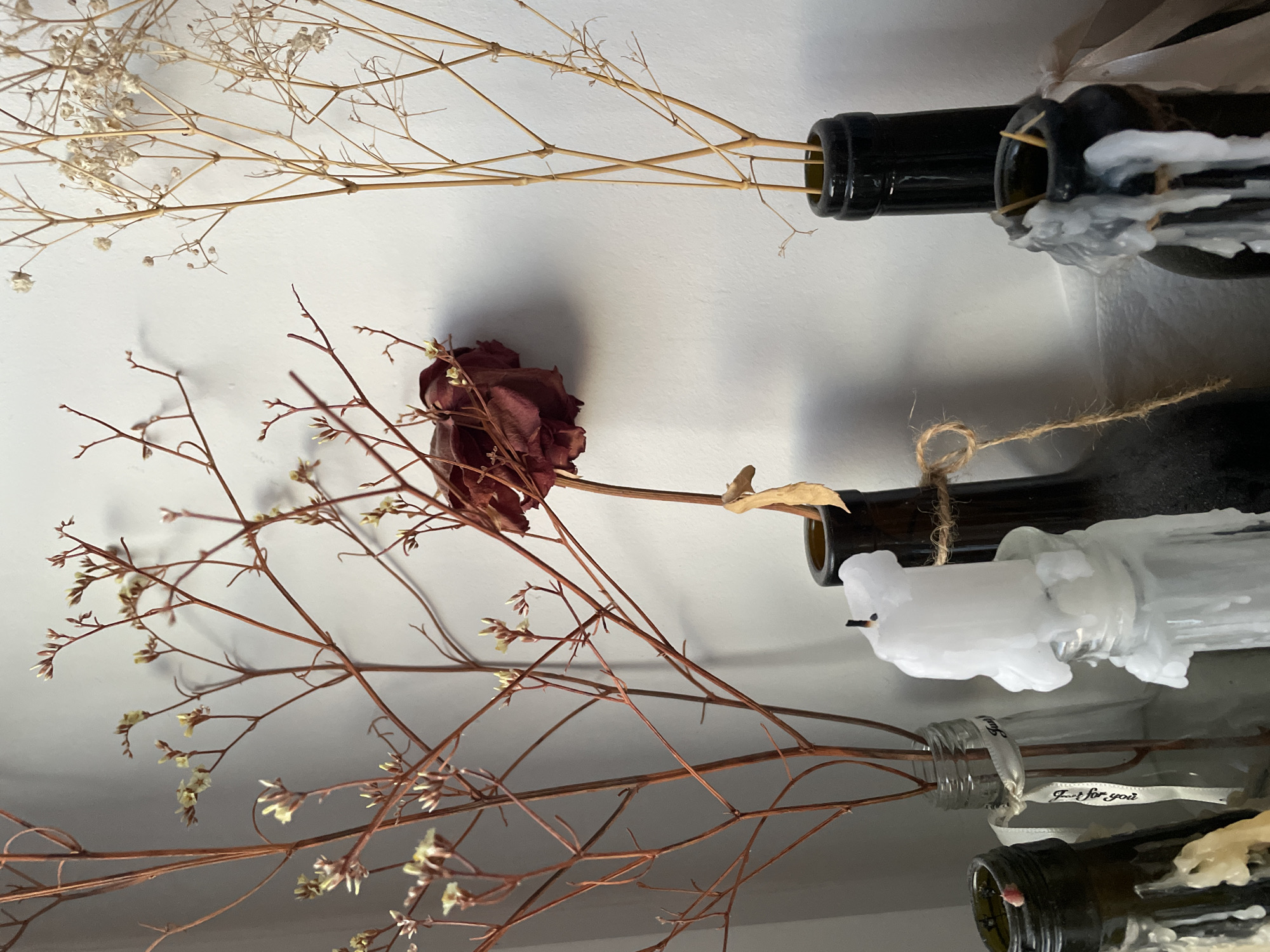 The image depicts a still life composition of dried flowers and candles in bottles, set against a plain white wall. The arrangement includes several dried floral branches, one with small yellow buds, and a single dried dark red rose. These are placed in various glass and dark-colored bottles. A white candle, partially melted and dripping wax, sits in one of the bottles. Another dark bottle is tied with twine. The lighting suggests an indoor environment, possibly a windowsill or shelf. The background is a smooth, unadorned white wall. No specific location in Hrodna, Belarus is identifiable from the image.