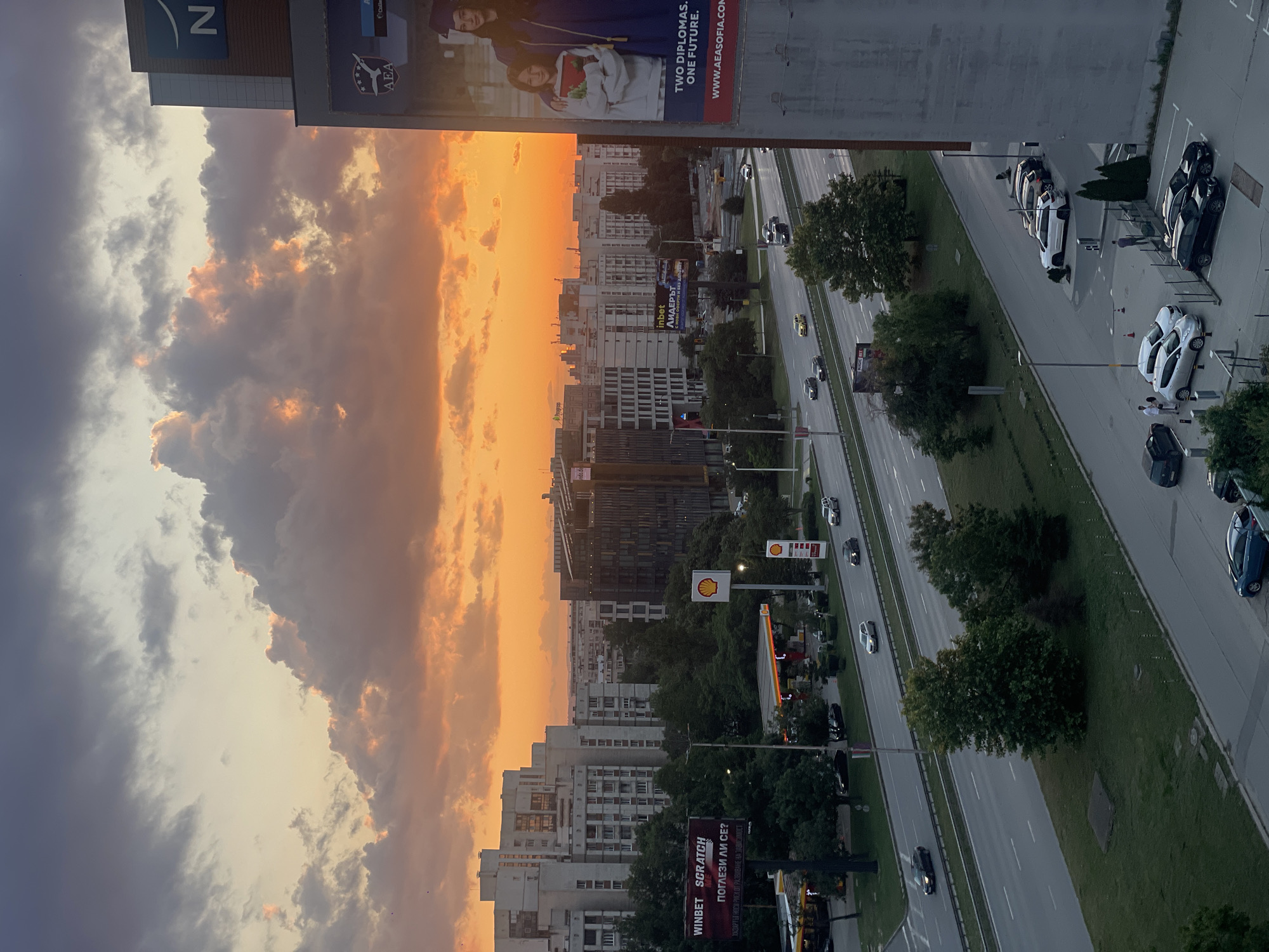 This image captures an urban landscape at sunset, characterized by an orange and yellow sky with numerous clouds, some illuminated by the setting sun and others appearing dark grey. The scene is dominated by a multi-lane highway running horizontally across the mid-ground, featuring an asphalt surface with visible lane markings and a central grassy median containing trees. Numerous vehicles, including sedans and SUVs of various colors (white, black, grey), are traveling on the highway in both directions.
On the left side of the highway, an active Shell gas station is visible, identifiable by its distinctive yellow and red canopy and branding, with fuel pumps and parked vehicles. Adjacent to it, a large billboard displays "WINBET SCRATCH ME" and Bulgarian text. Further into the background on the left, several large, multi-story residential buildings with grey facades and numerous windows stand prominently.
On the right side of the image, a large, modern building with a dark facade, possibly an office complex, is partially visible, featuring a large "N" logo near its top. A significant vertical billboard on the far right advertises "AEA SOFIA" (American English Academy Sofia), featuring images of two individuals, one in a graduation gown, and the text "TWO DIPLOMAS. ONE FUTURE." along with its website. Behind these structures, additional urban buildings extend into the horizon, silhouetted against the bright sky.
In the foreground, a parking lot and a street run parallel to the highway. Several cars are parked, including white, black, and blue models. A few individuals are present in this area; two people are standing near a white car, and another person is walking. Lush green trees are scattered throughout the scene, particularly along the highway median and in various green spaces. The overall impression is a bustling urban area during the twilight hours. The presence of the "AEA SOFIA" billboard confirms the location as the city of Sofia, Bulgaria.