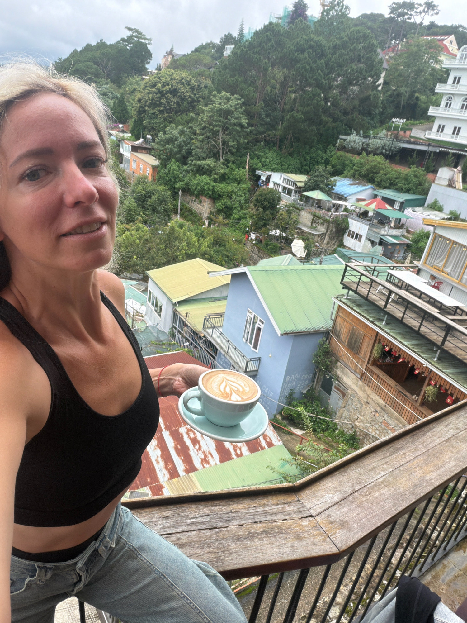 A blonde woman with fair skin and light-colored eyes is taking a selfie on a balcony, holding a cup of latte art coffee. She is wearing a black tank top and light blue jeans. The background shows a lush, green hillside filled with trees and buildings with colorful roofs, characteristic of the city of Da Lat, Vietnam. The buildings are a mix of architectural styles, with some appearing older and others more modern. The overall scene suggests a scenic overlook or a cafe with a view in Da Lat.