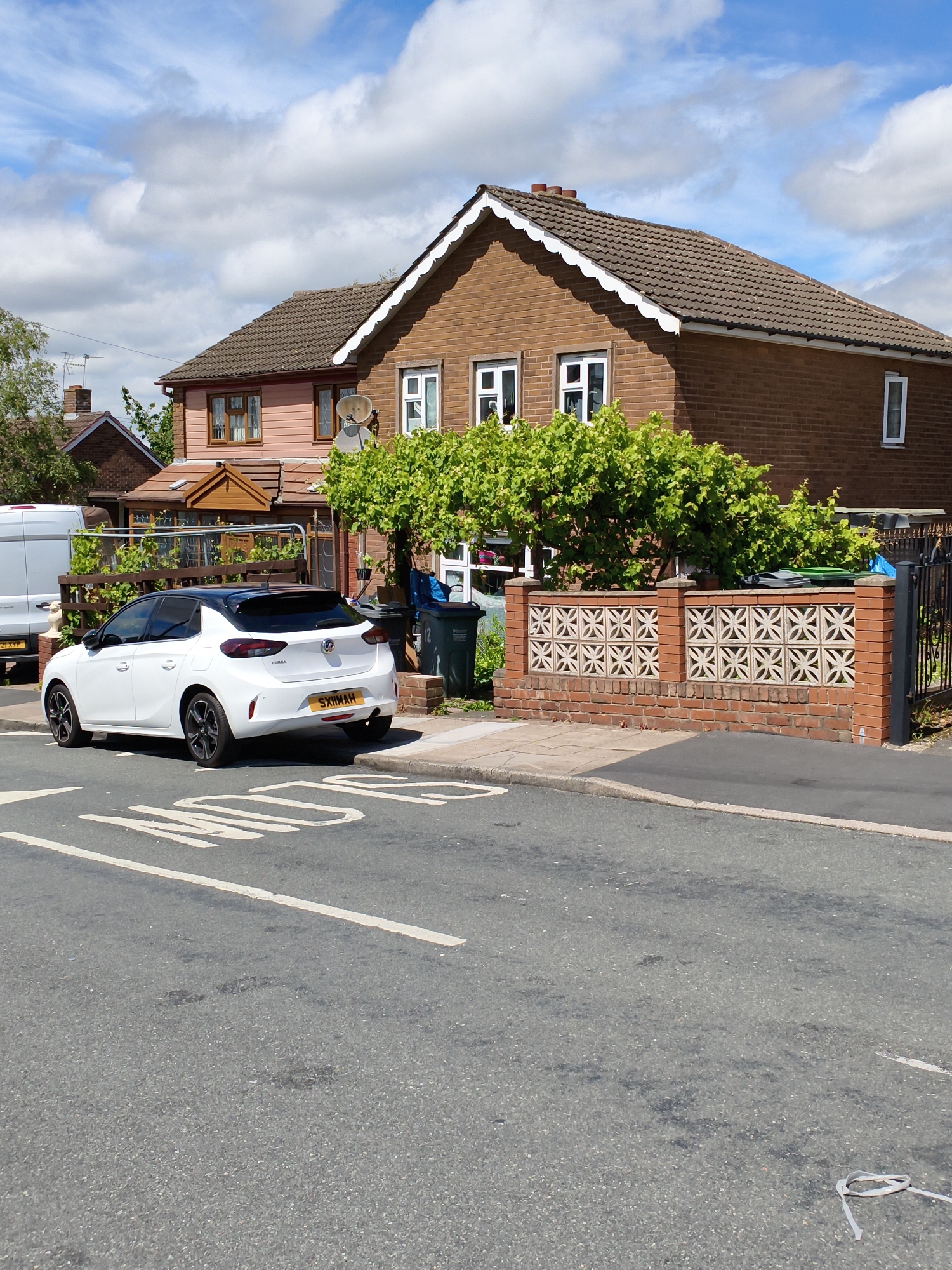 In Sandwell, United Kingdom, on a sunny day with scattered clouds, a white Vauxhall Corsa is parked on the left side of a residential street. The car, featuring a black roof and tinted windows, has a yellow license plate with the registration "SXIIMAH". A speed bump is visible on the asphalt road in the foreground.
Behind the car, a brick house with a brown tiled roof and white decorative trim stands. The house has multiple windows, some with white frames and others with brown frames. A porch area with wooden supports is attached to the front. Adjacent to the house, a low brick wall with decorative concrete panels separates the property from the street. Green trees and foliage grow behind the wall and around the house. Two dark green wheelie bins are positioned near the property's entrance.
To the left of the house, part of a white van is visible, with a license plate that reads "ZI XYP". The environment appears to be a suburban residential area.