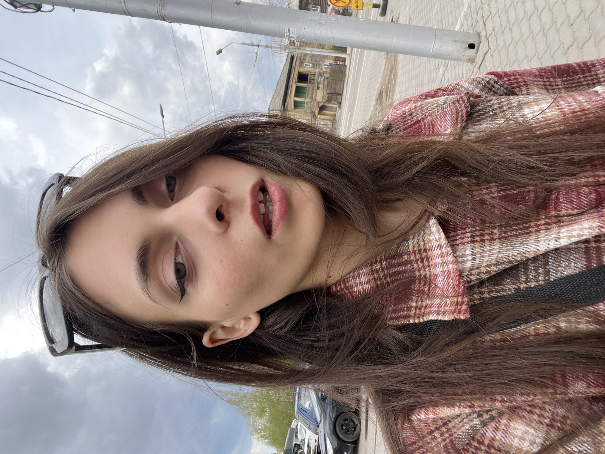A selfie of a young woman with long brown hair wearing a plaid shirt. She's standing outside on a street with a power pole visible in the background. Across the street is an old, dilapidated two-story building with dark windows. The sky is overcast. There are parked cars and trees in the lower part of the image, suggesting a street scene. A sign showing "No Stop" is visible in the background. Based on the style of the old wooden buildings and street signage, this image appears to be taken in Krasnoyarsk, Russia.