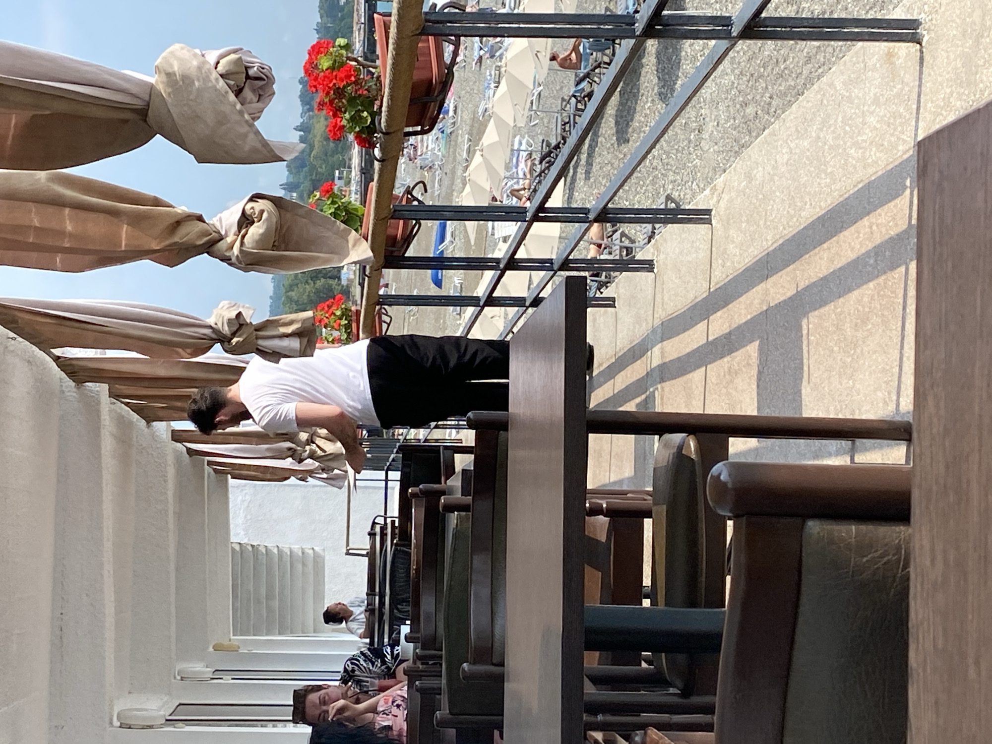 The image depicts an outdoor seating area of a cafe or restaurant located on the waterfront in Chabkwara, Abkhazia. The foreground shows several dark wooden tables and chairs, some of which are partially obscured. In the middle ground, a man in a white t-shirt and black pants is standing and appears to be holding a tray, possibly serving customers. To his left, two women are seated at a table, one of whom is looking down and holding her face. In the background, beyond a metal railing, there is a beach with numerous lounge chairs and umbrellas. The sea is visible further out, with a distant shoreline and trees. Red flowers in planters are positioned along the railing, adding a touch of color to the scene. The overall impression is one of a relaxed, sunny day at a seaside establishment.
