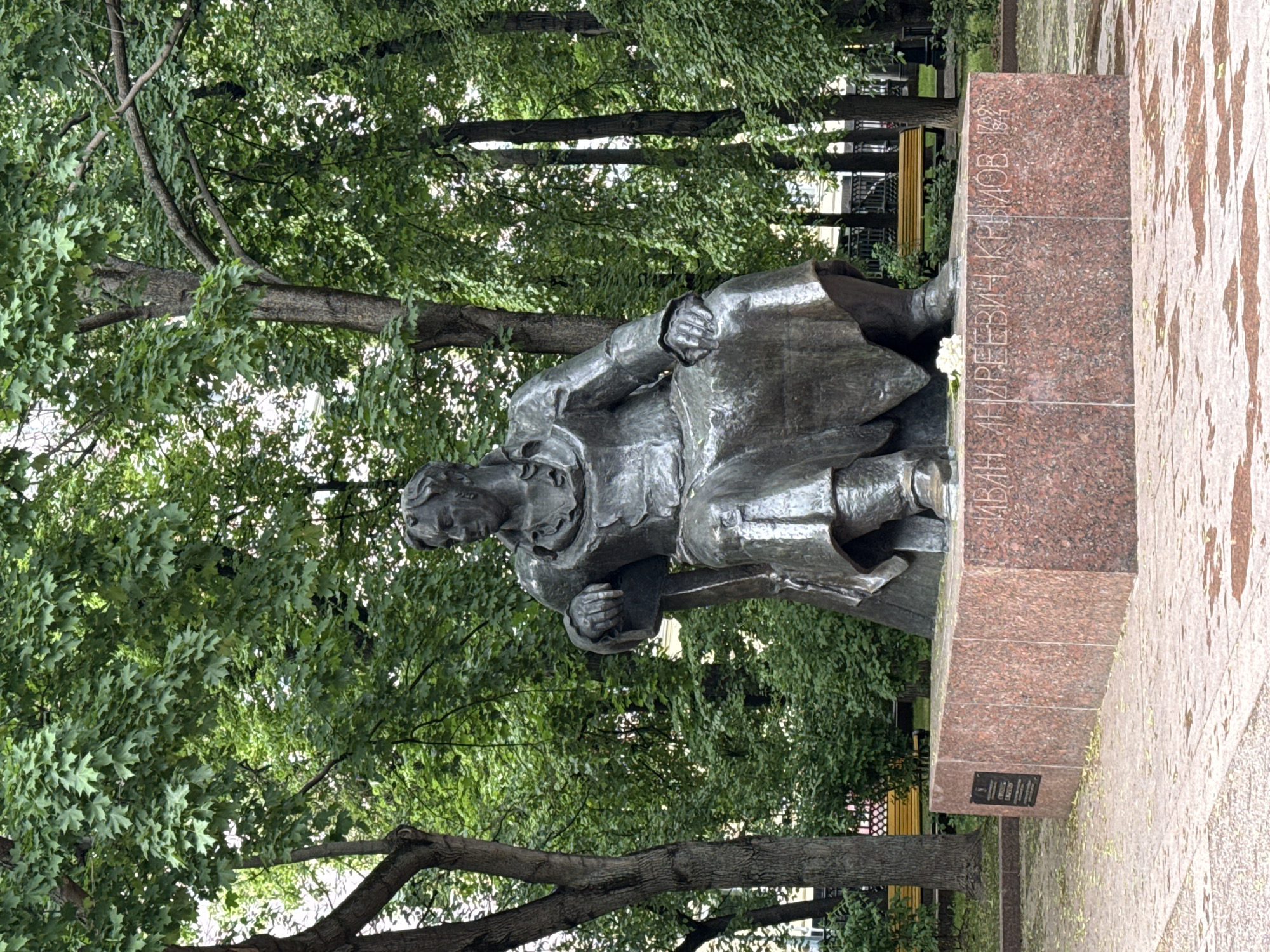 The image captures a statue of Ivan Andreevich Krylov, a Russian fabulist, located in Moscow, Russia. The bronze statue depicts Krylov in a seated position, seemingly deep in thought, with a book in his hands. It is set against a backdrop of lush green trees, suggesting a park or garden setting. The statue stands on a granite pedestal inscribed with his name and dates. The surrounding area shows a paved surface, possibly a pathway within the park.